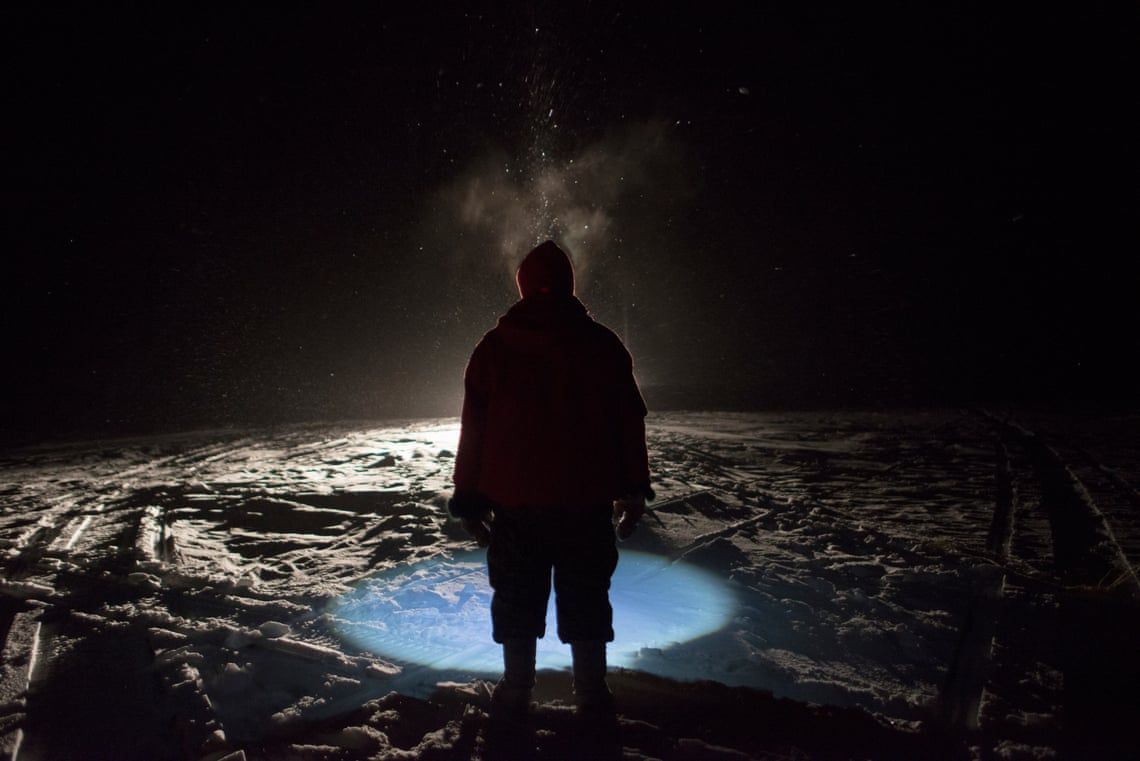 A Canadian ranger, illuminated by a snowmobile and his headlamp during a snowstorm, helps pull a net using under-ice fishing techniques for Arctic char during a patrol on King William Island, Nunavut. Canadian rangers are a volunteer, community-based military unit. In the Arctic they are mostly Inuit. Image by Louie Palu / Zuma Press. Canada, undated. 