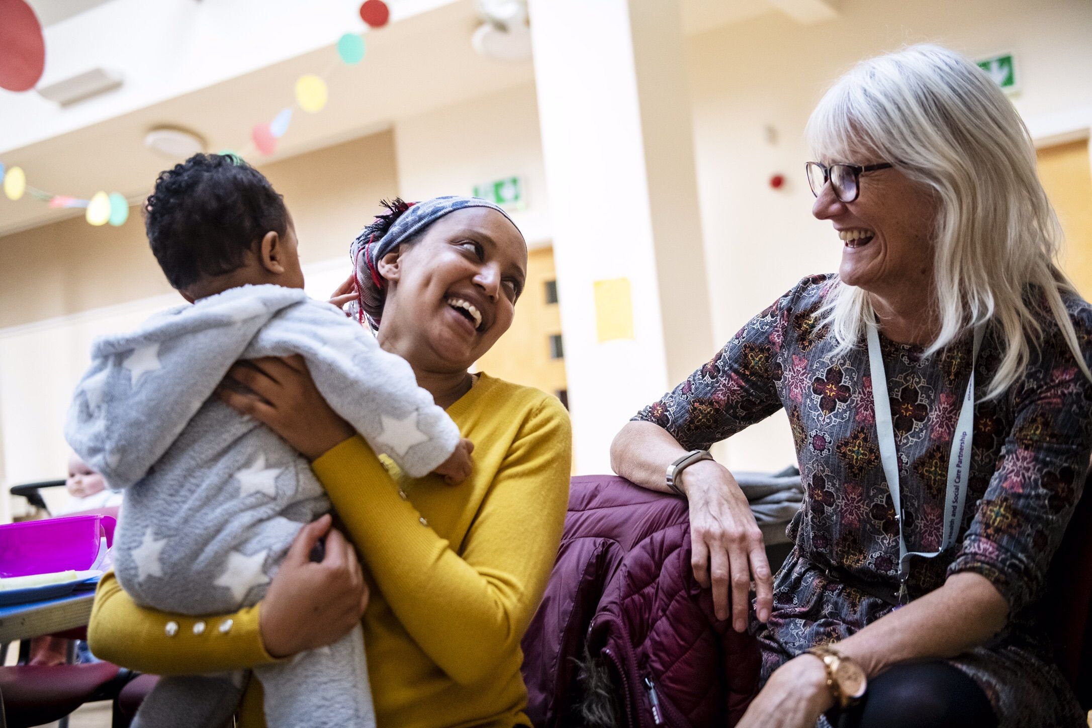 Eliad Abrha, 7 months, left,  is held by his mother Hanna Abrha, of Parkhead, a neighborhood of Glasgow, Scotland, center, as she speaks and jokes with Janet Tobin, health improvement lead with the National Health Service, right, at Cafe Stork, Tuesday, Oct. 21, 2019, at Parkhead Congregational Church in Glasgow. Cafe Stork is a free networking lunch for families in the disadvantaged East End who are expecting or new parents. Image by Michael M. Santiago. United Kingdom, 2019.