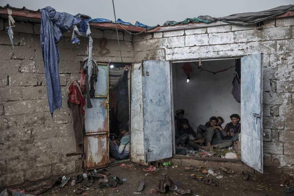 Yemenis, right, chew qat, while Ethiopian migrants, left, take shelter in a small shack at a qat market in Dhale province in Yemen. This is one of the stops where migrants take shelter to continue their journey to Saudi Arabia, and an active frontline between militiamen backed by the Saudi-led coalition and Houthi rebels only a few hundred meters (yards) away. Image by Nariman El-Mofty. Yemen, 2019.