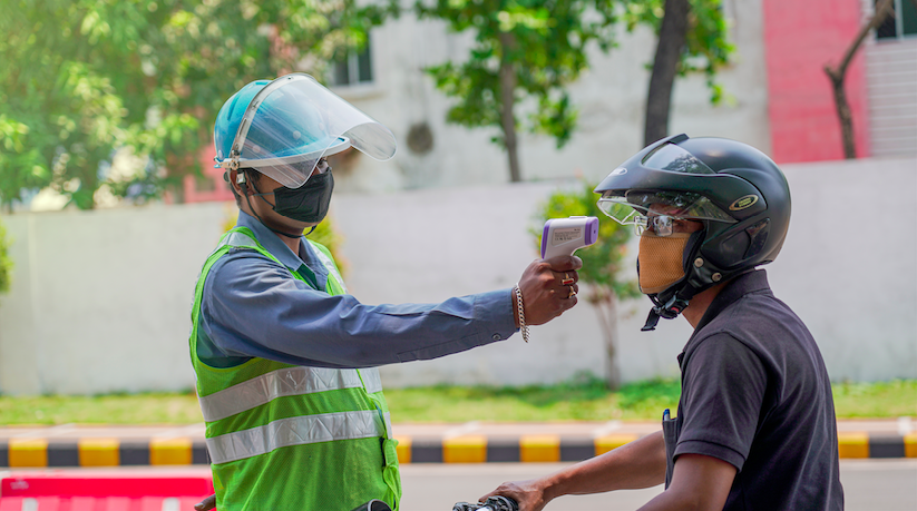 A medical worker in Jamshedpur checks the body temperature of a biker with an infrared thermometer gun for COVID-19 symptoms. Image by ArnavIG / Shutterstock. India, 2020.