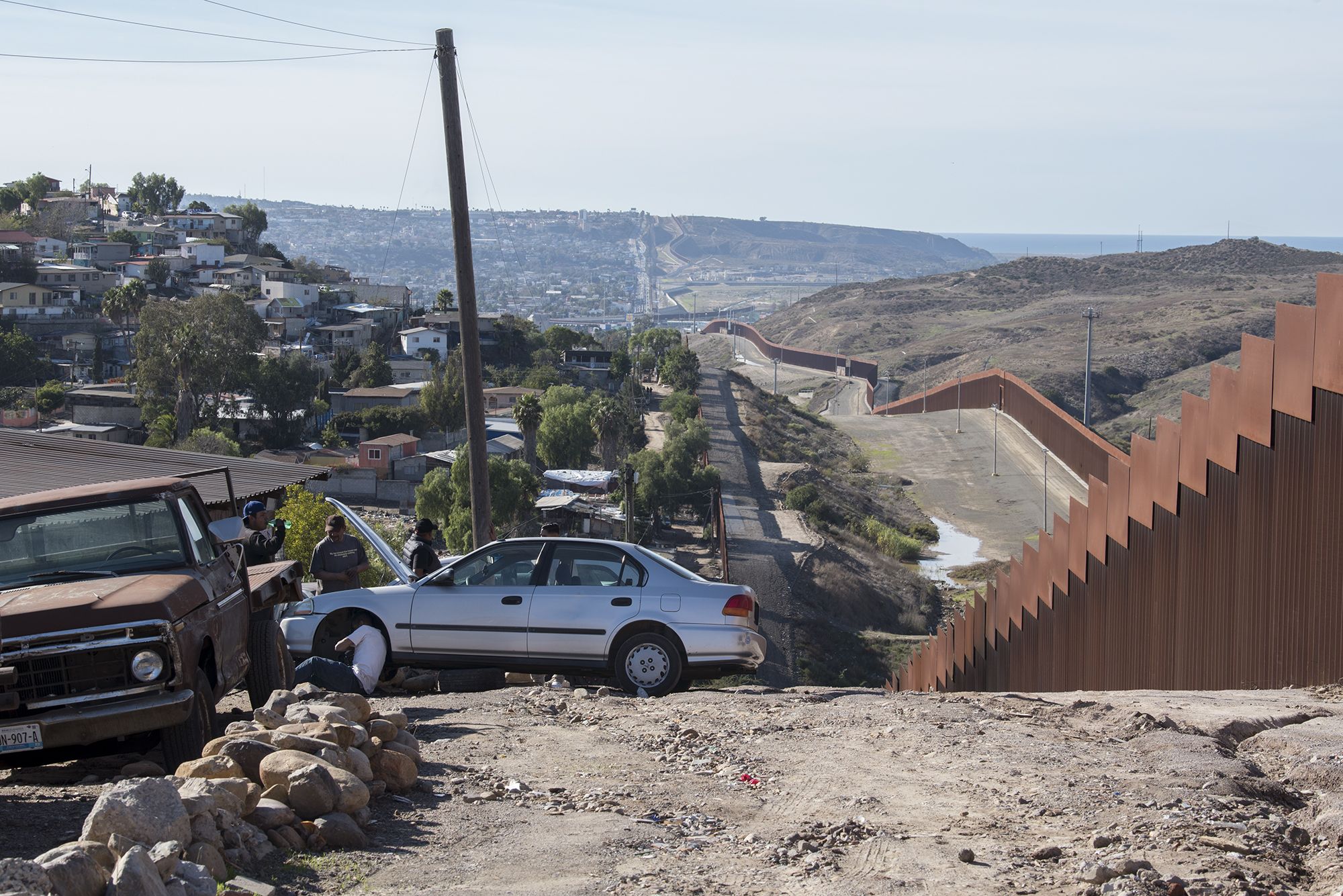 Men gather to fix a car in the Libertad Parte Alta neighborhood of Tijuana, Mexico, left, as the border wall stretches on for miles nearby. Image by Amanda Cowan. Mexico, 2019.