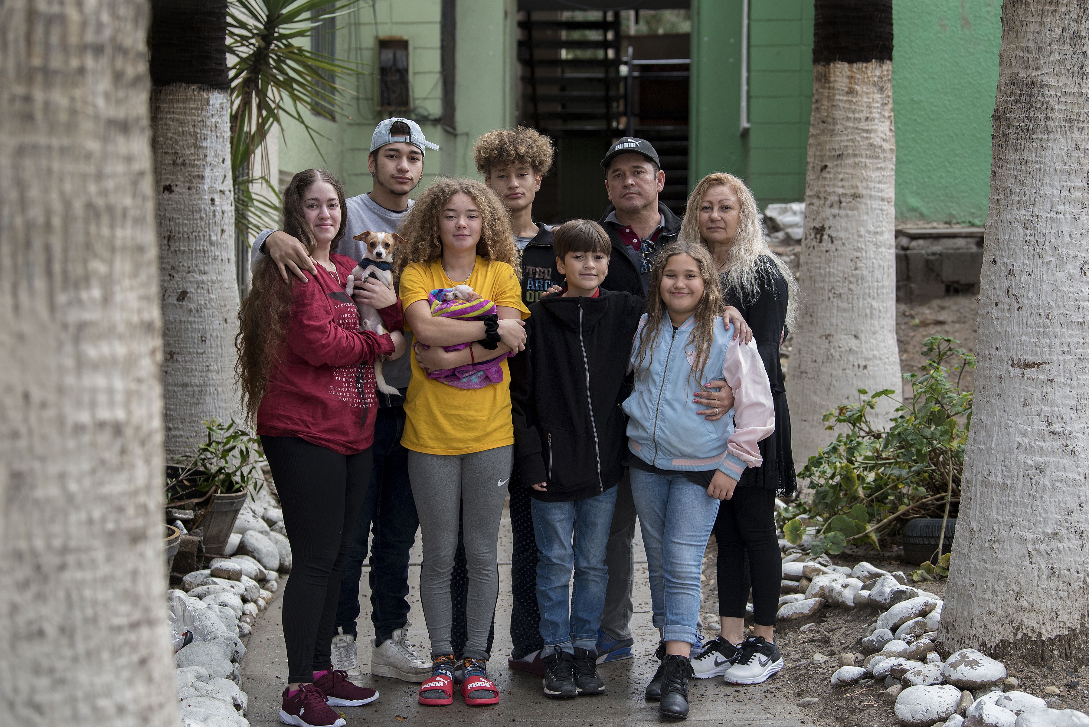 The Flores family pauses for a portrait outside Ramon’s apartment complex in Tijuana, Mexico. Family members are, clockwise from left, Leslie, 23, Lennes, 18, Raymond, 15, Ramon, 46, Enedis, 54, Rayma, 10, Edward, 12, and Kennedy, 16. The dogs are Canelo, left, and Bambi, wrapped in the blanket. Image by Amanda Cowan. Mexico, 2019.