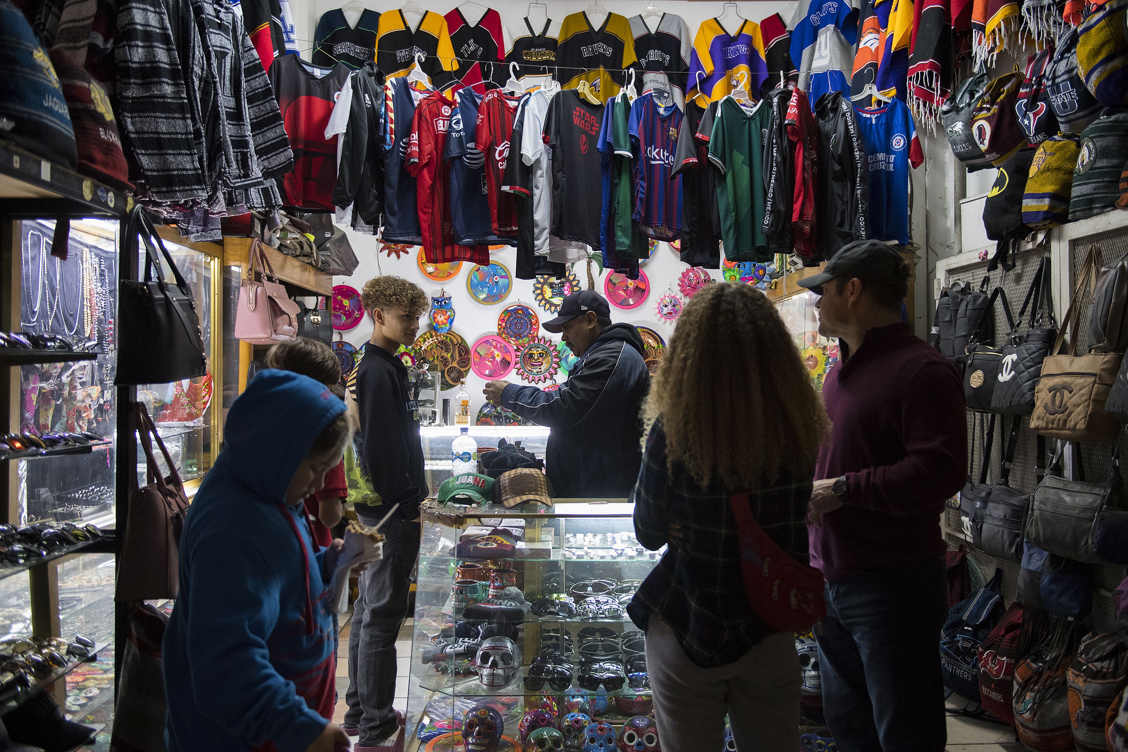 Raymond Flores looks to his father, Ramon, right, for approval as he shops for his birthday present with his siblings along Avenida Revolucion in Tijuana, Mexico. The chain costs 750 pesos, which is approximately $38. Ramon, who makes around $110 a week, said he wanted him to have the gift. "When I get the opportunity to give them something they want, I feel good," he said. Image by Amanda Cowan. Mexico, 2019.