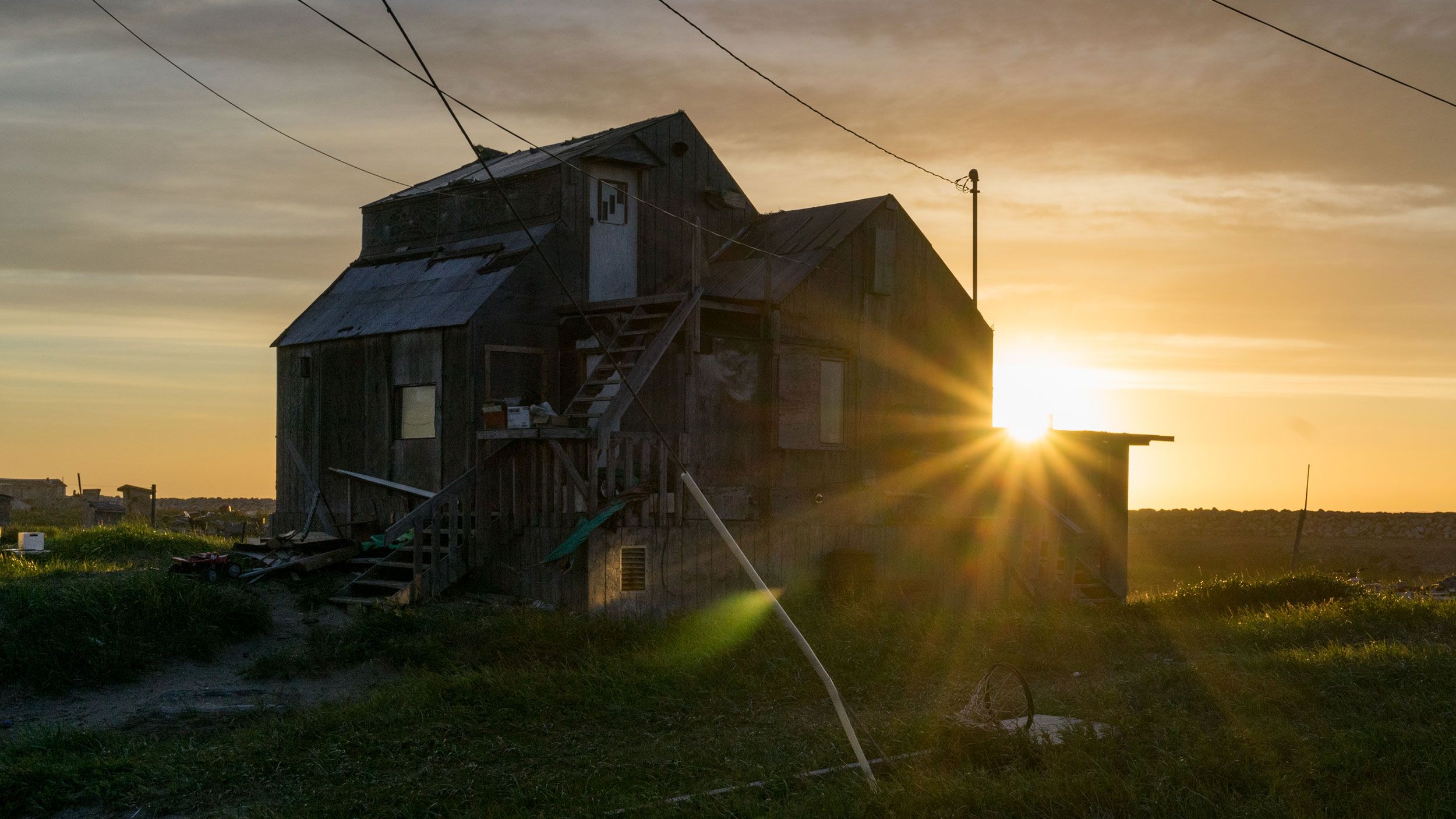 Rays of sunlight spill over a  Shishmaref home as the midnight sun sets over the Chuckchi Sea. Image courtesy of Nick Mott. United States, 2018.