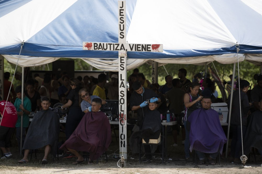 In this Oct. 11, 2019 photo, migrants get free haircuts by volunteers at a migrant camp near a legal port of entry bridge in Matamoros, Mexico. Tamaulipas became a bloody emblem of the problem of violence against migrants in 2010 when 72 migrants were found slain at a ranch in San Fernando, and a year later when the bodies of 193 migrants were found in the same area in clandestine mass graves, apparently murdered by a cartel to damage a rival’s people-smuggling business. Image by Fernando Llano. Mexico, 2019.