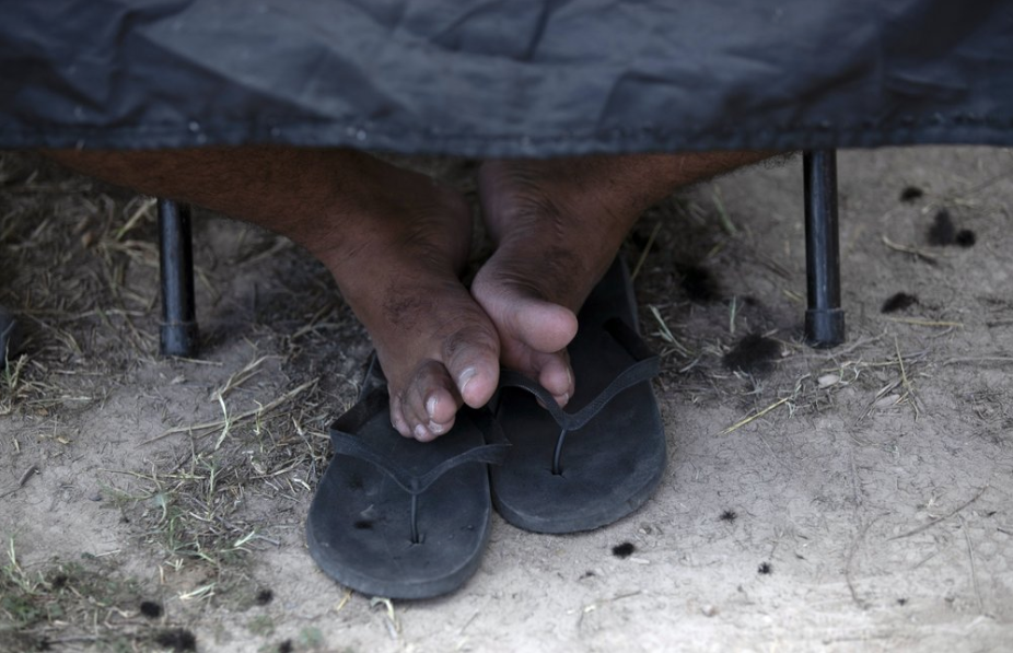 In this Oct. 11, 2019 photo, the feet of a migrant boy rest on his sandals as he sits still for a haircut by a volunteer at a migrant camp near the legal port of entry bridge that connects Matamoros, Mexico with Brownsville, Texas. In years past, migrants moved quickly through this violent territory on their way to the United States. Now, due to Trump administration policies, they remain in Mexico's Tamaulipas state for weeks and sometimes months as they await their U.S. court dates, often in the hands of the gangsters who hold the area in a vice-like grip. Image by Fernando Llano. Mexico, 2019.