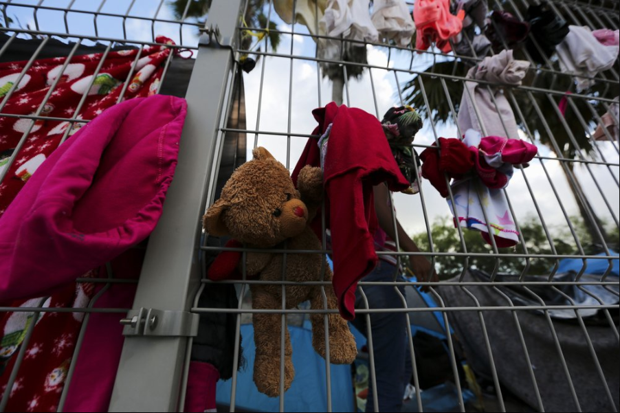 In this Oct. 11, 2019 photo, a stuffed teddy bear hangs in a fence amid clothing at a migrant camp set up near a legal port of entry bridge that connects Matamoros, Mexico with Brownsville, Texas. There’s not enough space for everyone at the shelters, so many rent rooms, ranging from $35 per person per month for a spot in a cramped five-person bedroom in a seedy area, to $300-$500 for a more secure home. But nowhere is truly safe. Image by Fernando Llano. Mexico, 2019.