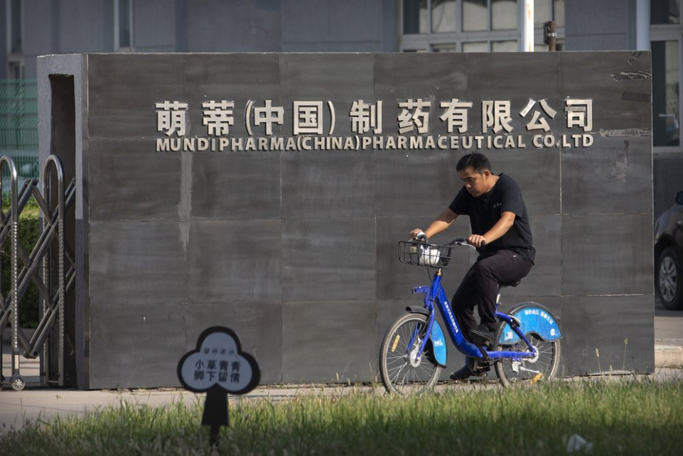A man rides a bicycle past the entrance gate of a Mundipharma facility in an industrial park on the outskirts of Beijing, China on Sept. 27, 2019. As the Sackler's U.S. empire collapses, Mundipharma, which is also owned by the family, is using the same tactics to sell opioids in China. Image by Mark Schiefelbein / AP Photo. China, 2019.