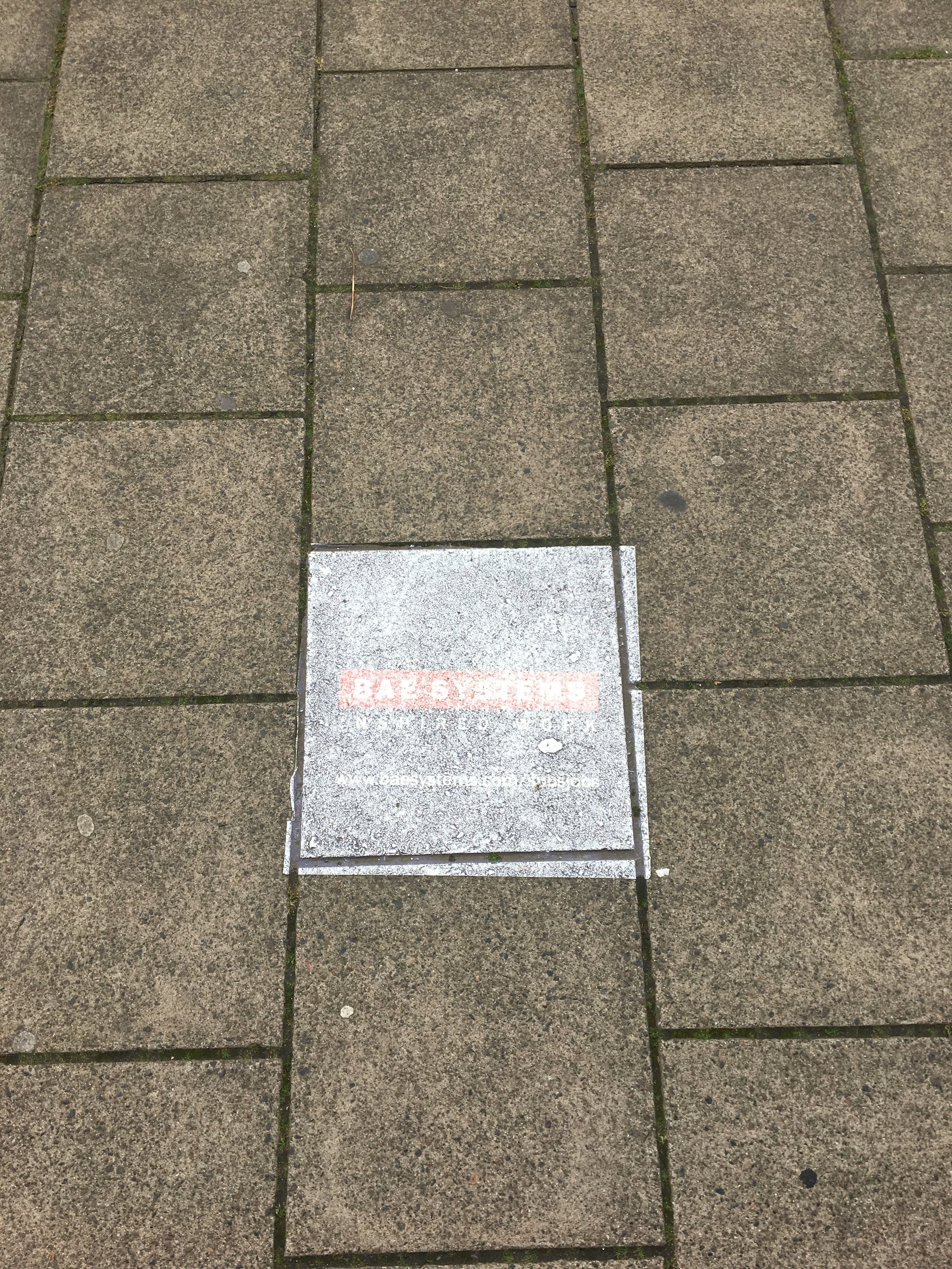 BAE Systems has penetrated every part of Barrow-in-Furness. Here its logo has been sprayed Banksy-style on the pavement in central Barrow. Image by Matt Kennard. United Kingdom, 2017.
