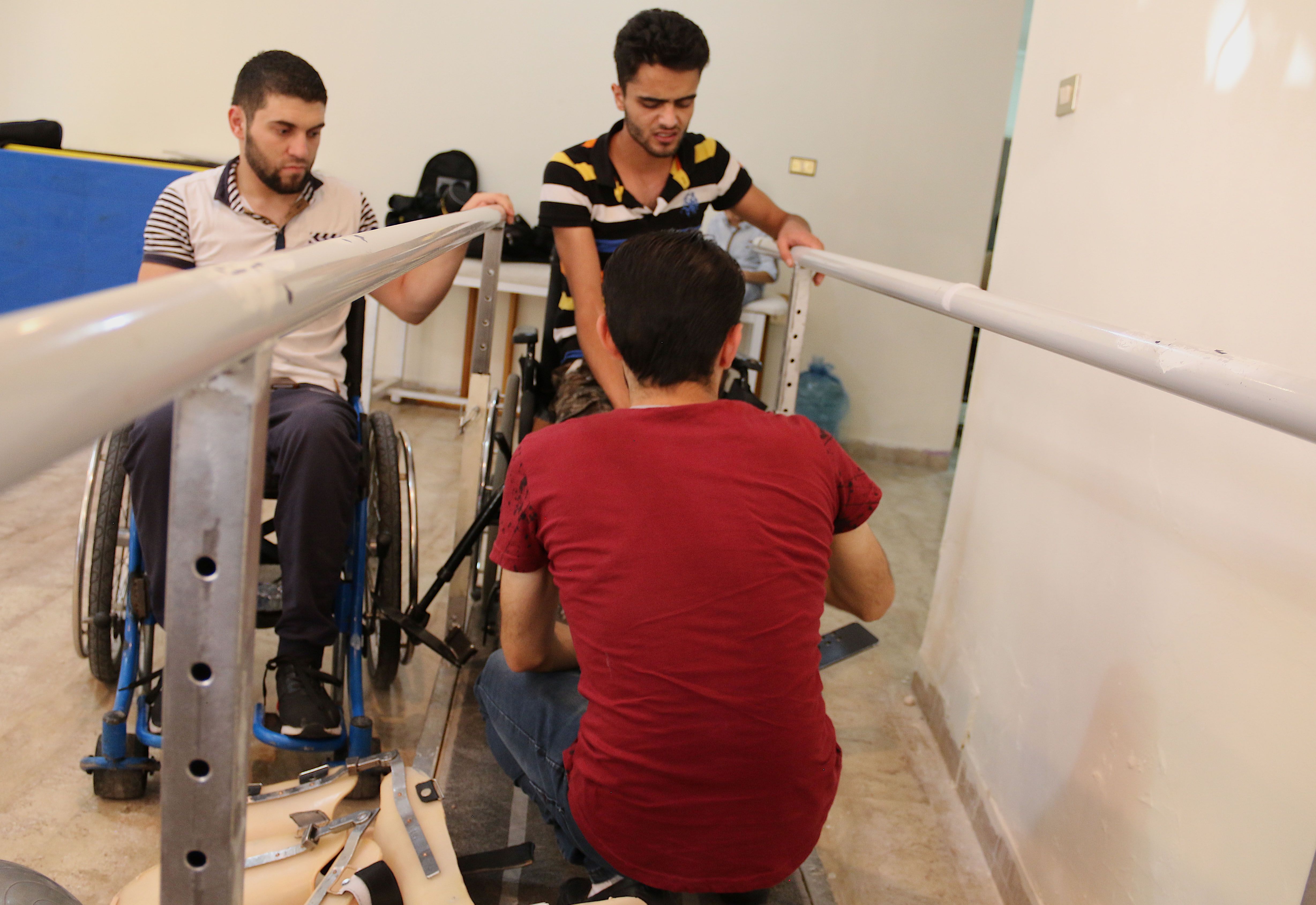 Obada Alasmi watches as a trainer helps Mohammed Ayyash lift his legs. Ayyash was paralyzed in an attack in Syria. Image by Sawsan Morrar. Jordan, 2017.