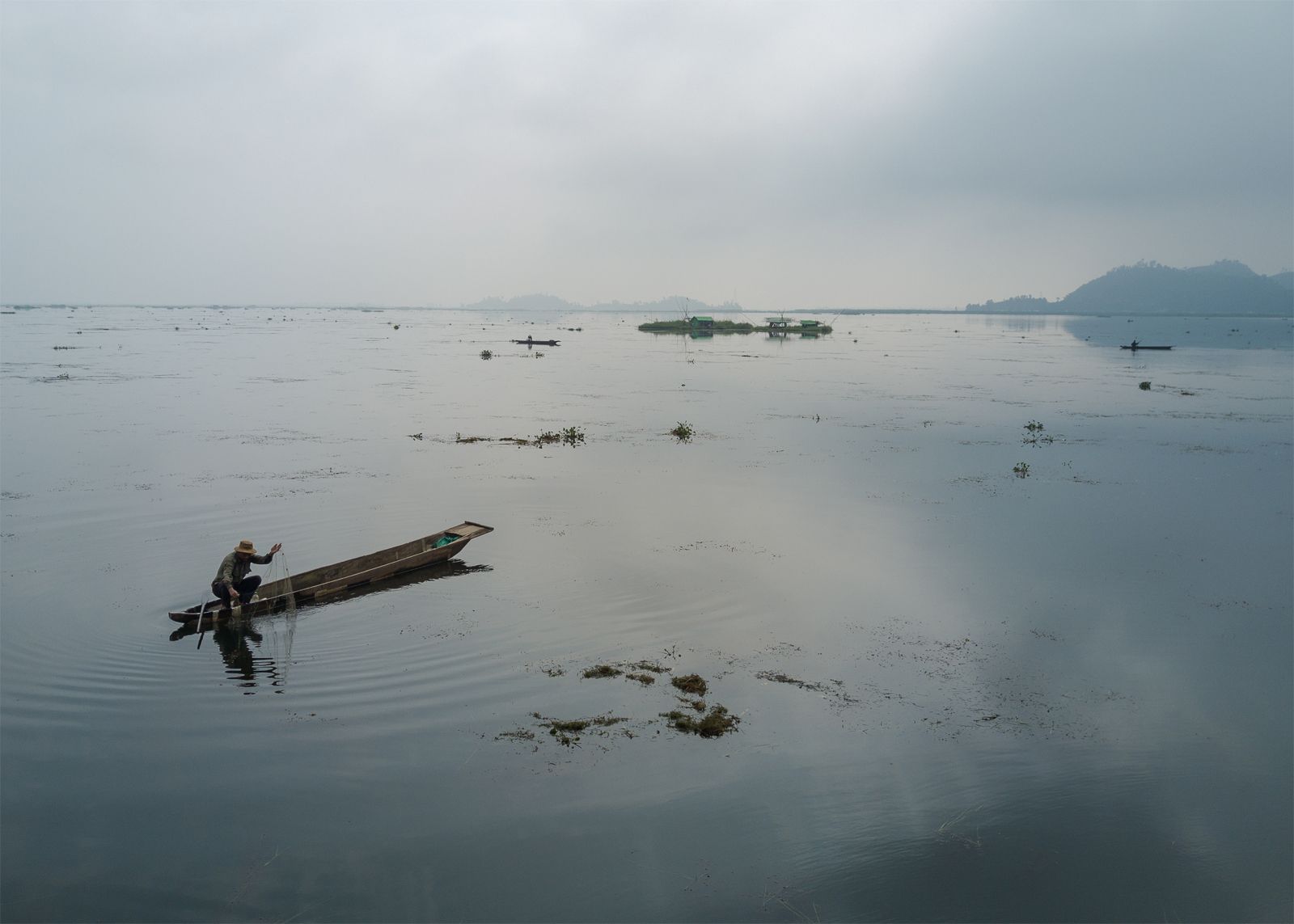 A Meitie fisherman fishes near phumshangs (floating huts) in Loktak Lake, Manipur. The construction of the Ithai Dam flooded over 80,000 hectares of farm and pasture land in the vicinity of the Loktak wetland. The Loktak Development Authority has been dredging the phumdis (floating biomass) and has cleared over 800 Phumshangs in the name of lake clean-up for efficient power generation. Image by Neeta Satam. India, 2017.