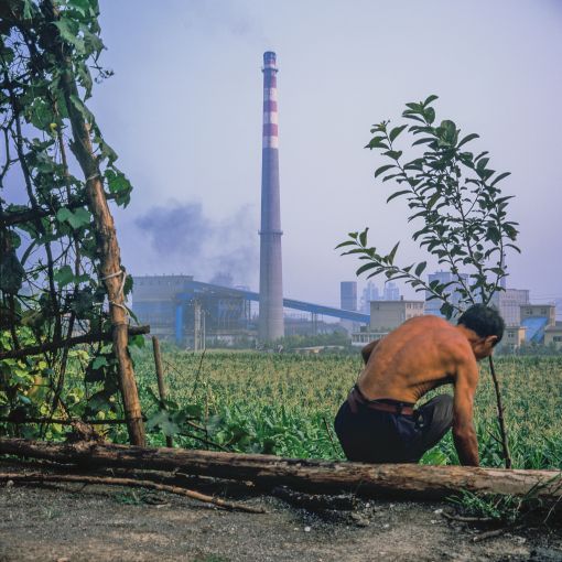 A resident of Sihoupo village, about 40 km southwest of downtown Handan, leaps down into his vegetable plot right outside his home to work the fields. The coking plant in the background — a branch of the Hansteel works which dominates Handan city — is less than 100 m from the houses in this village. Day and night, the factory emits steam and smoke in cycles — grey, black or yellow — and constantly beeps like an electrocardiogram machine. Depending on the direction of the winds, the smoke sometimes blows low right across these farm plots where villagers grow corn and vegetables.  Image by Sim Chi Yin. China, 2013.