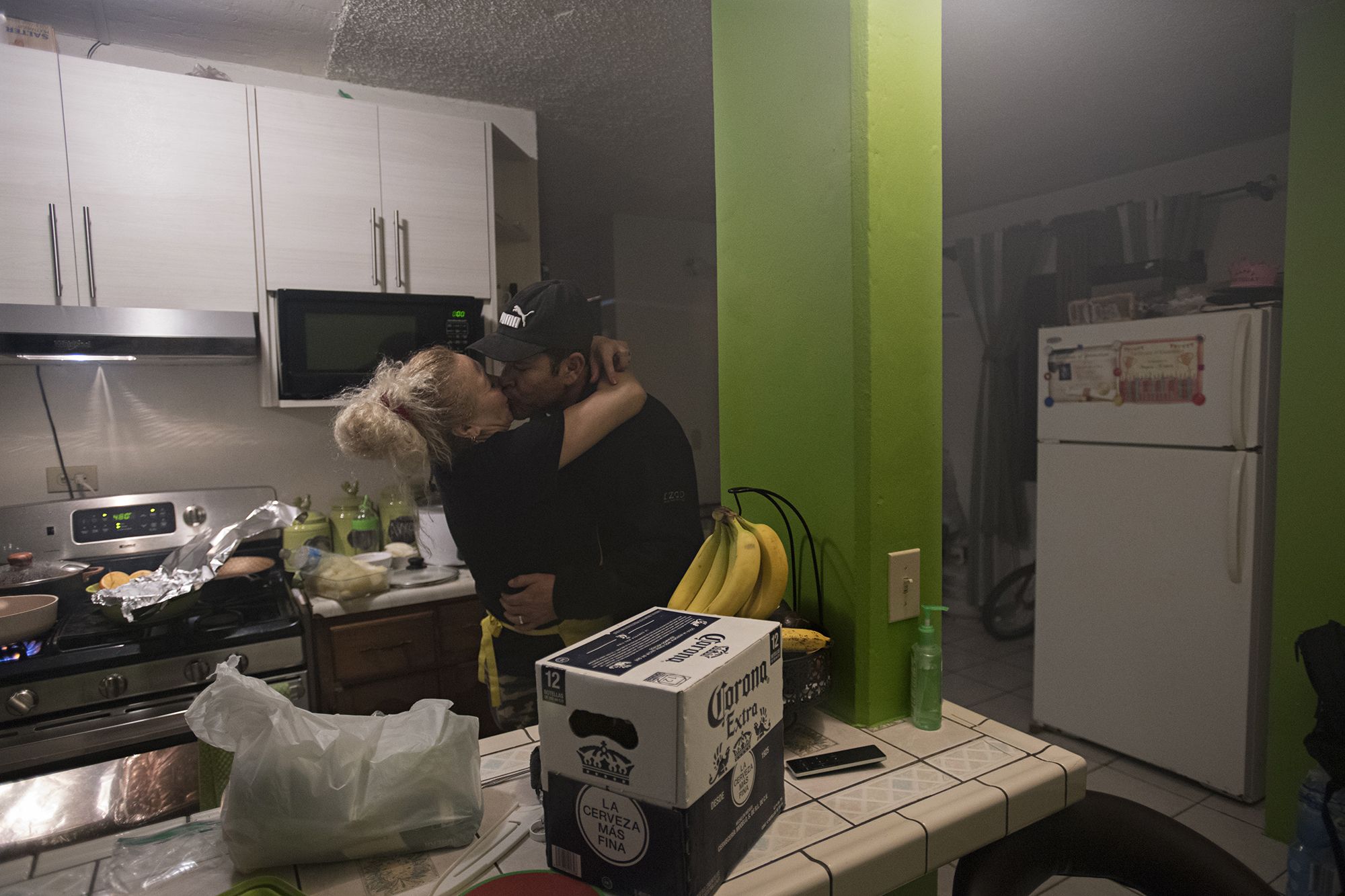 Enedis Flores welcomes home her husband, Ramon, from work as she cooks Thanksgiving dinner at his apartment in Tijuana, Mexico. Ramon, who works as a carpenter, built custom cabinets and painted the walls of the apartment to make it feel more like home for his family. Image by Amanda Cowan. Mexico, 2019.