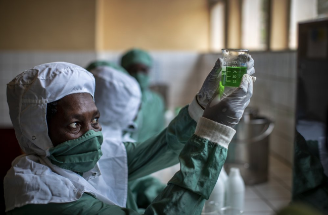In this photo taken Thursday, Nov. 7, 2019, workers wearing protective clothing to protect from the effects of the drug and to prevent contamination, make liquid morphine from powder which is dyed green as a color-code to indicate the strength, at the Pharmaceutical Laboratory of Rwanda in Butare, Rwanda. While people in rich countries are dying from overuse of prescription painkillers, people in Rwanda and other poor countries are suffering from a lack of them, but Rwanda has come up with a solution to its pain crisis - it's morphine, which costs just pennies to produce and is delivered to households across the country by public health workers. Image by Ben Curtis / AP Photo. Rwanda, 2019.