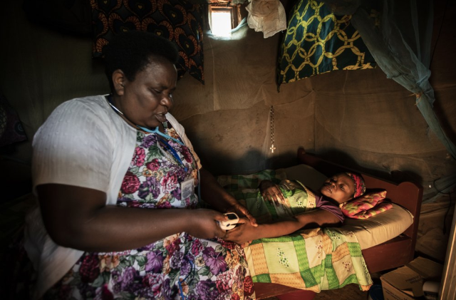 In this photo taken Tuesday, Nov. 5, 2019, palliative care nurse Madeleine Mukantagara, 56, left, uses a pulse-oximeter to check on the health of Vestine Uwizeyimana, 22, right, who has spinal degenerative disease and is taking oral liquid morphine for her pain, at her home in the village of Bushekeli, near Kibogora, in western Rwanda. While people in rich countries are dying from overuse of prescription painkillers, people in Rwanda and other poor countries are suffering from a lack of them, but Rwanda has come up with a solution to its pain crisis - it's morphine, which costs just pennies to produce and is delivered to households across the country by public health workers. Image by Ben Curtis / AP Photo. Rwanda, 2019.