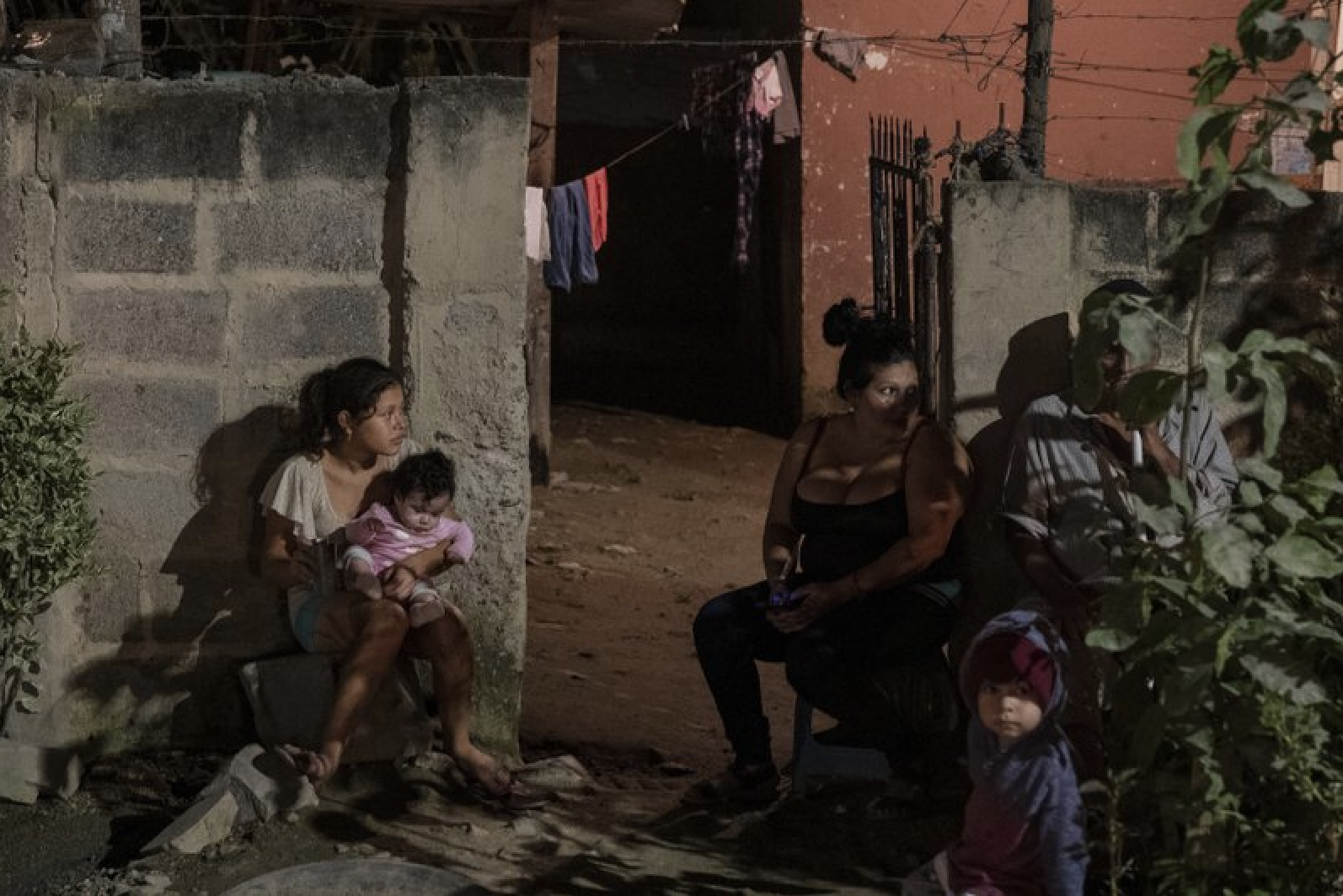 A family sits outside their home as forensic workers investigate a body at a crime scene in the Rivera Hernandez neighborhood of San Pedro Sula, Honduras on Nov. 30, 2019. MS-13. Mara 18. Los Vatos Locos _ The Crazy Guys. The gangs' shifting lines of control dodge and weave through Rivera Hernandez, a place where even the police are afraid. Image by Moises Castillo/ AP Photo. Honduras, 2019.