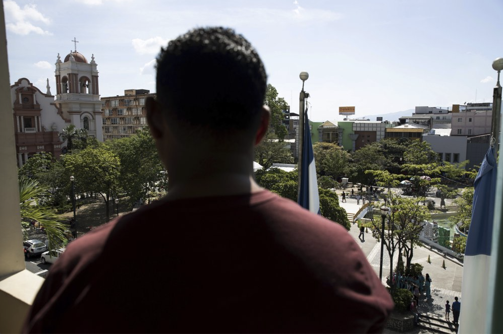 A Honduran man deported from the United States looks over the central park of San Pedro Sula, Honduras on Dec. 1, 2019. As he was walking in the area in November, a man stepped toward him, fired one shot from a pistol, and fled. He says he and his relatives have been hunted for more than 20 years by a powerful criminal family from his small hometown. "I've spent my whole life running. … One day they are going to get me." Image by Moises Castillo/ AP Photo. Honduras, 2019.