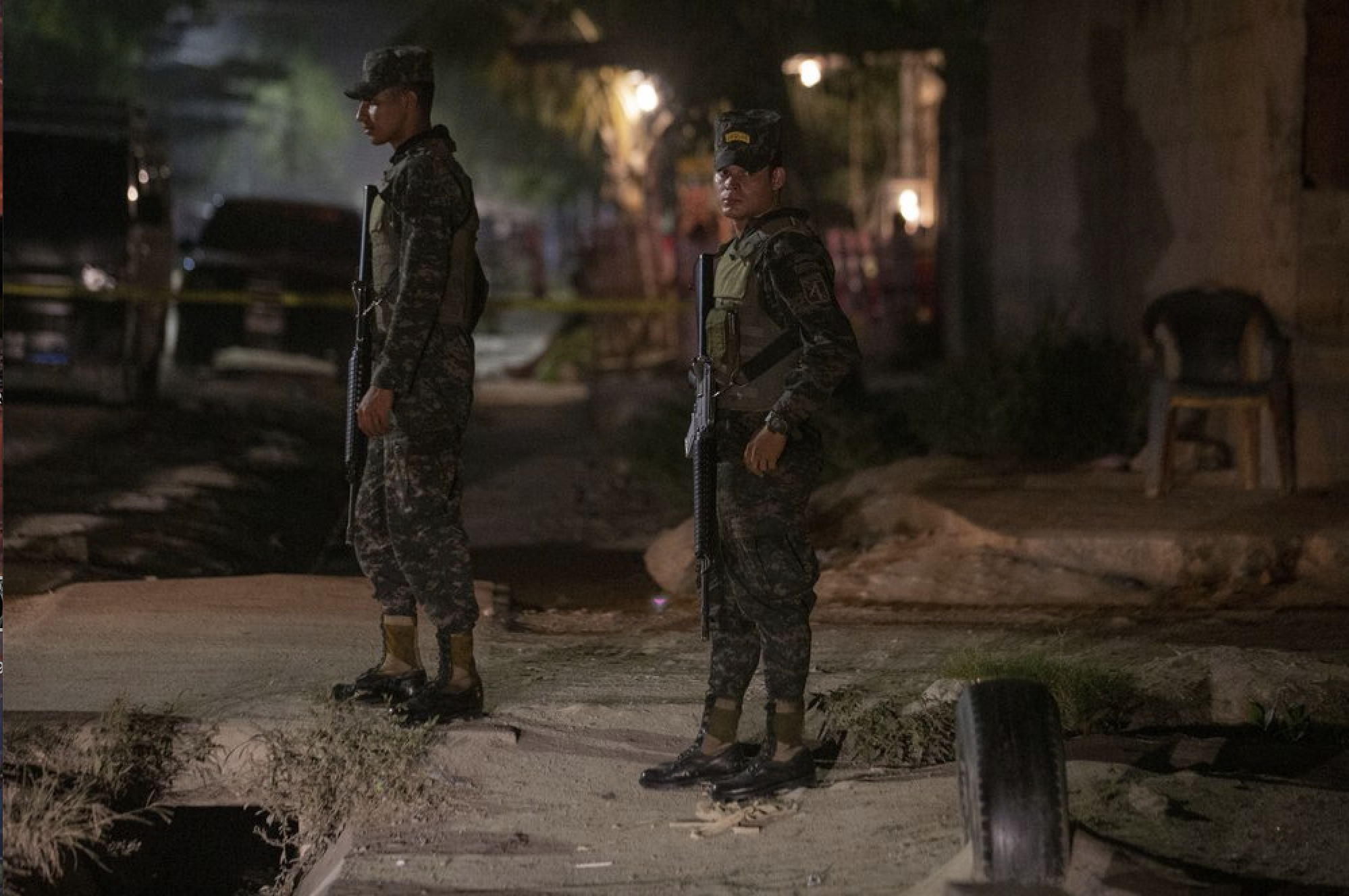 In this Nov. 30, 2019 photo, soldiers stand guard near a crime scene as forensic workers inspect a body in the Rivera Hernandez neighborhood of San Pedro Sula, Honduras. There are police stations in these neighborhoods, but everyone knows who is in charge. The gangs monitor the streets, the police patrols and rival gangs using a complex network of young boys who work in shifts around the clock and report anything suspicious. Image by Moises Castillo/ AP Photo. Honduras, 2019.
