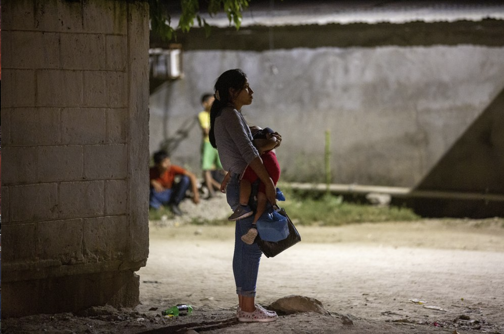 A woman holds a child near a crime scene as forensic workers inspect a body on the street in the Rivera Hernandez neighborhood of San Pedro Sula, Honduras, on Nov. 30, 2019. Image by Moises Castillo/ AP Photo. Honduras, 2019.