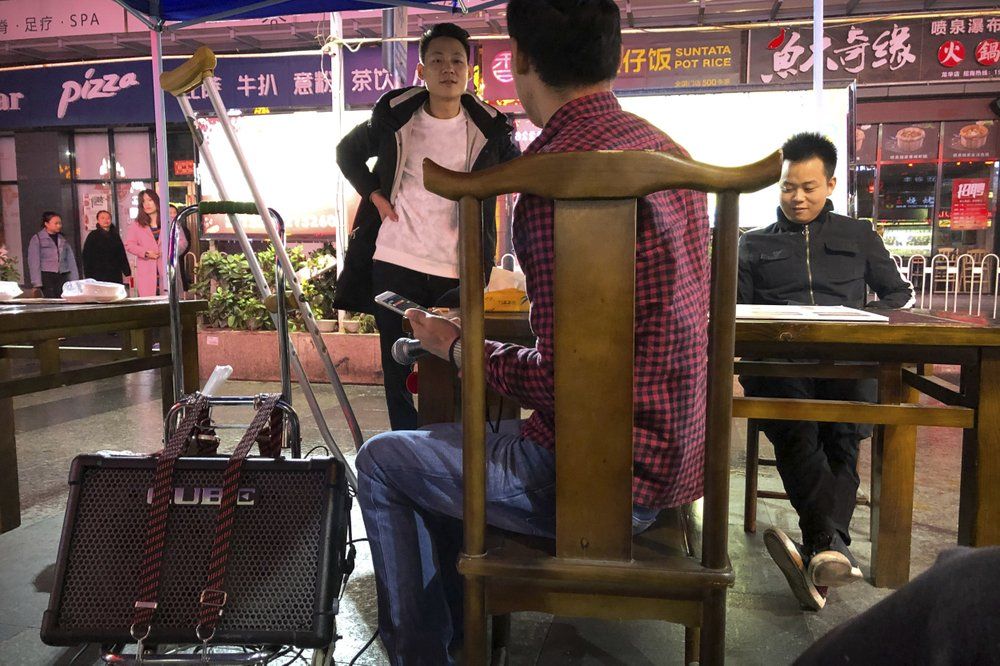 In this Dec. 6, 2019, photo, passers-by talk to Wu Yi, who has struggled with Oxycontin abuse, as he waits for a break in the rain while making his rounds to sing songs for money at all night-restaurants and clubs in Shenzhen in southern China's Guangdong Province. Officially, pain pill abuse is an American problem, not a Chinese one. But people in China have fallen into opioid abuse the same way many Americans did, through a doctor's prescription. And despite China's strict regulations, online trafficking networks, which facilitated the spread of opioids in the U.S., also exist in China. Image by Mark Schiefelbein/ AP Photo. China, 2019.