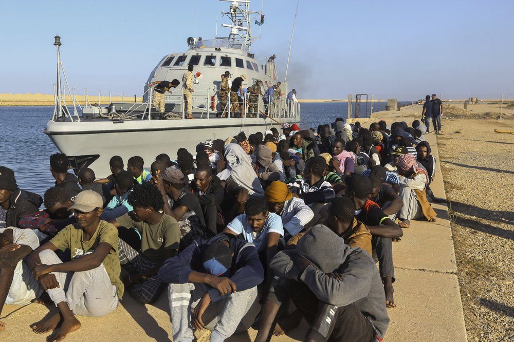 In this Tuesday, Oct. 1, 2019 file photo, rescued migrants are seated next to a coast guard boat in the city of Khoms, Libya, around 120 kilometers (75 miles) east of Tripoli. When millions of euros started flowing from the European Union into Libya to slow the tide of migrants crossing the Mediterranean, the money came with EU promises to improve detention centers notorious for abuse and to stop human trafficking. That hasn’t happened. Image by Hazem Ahmed/ AP Photo. Libya, 2019.