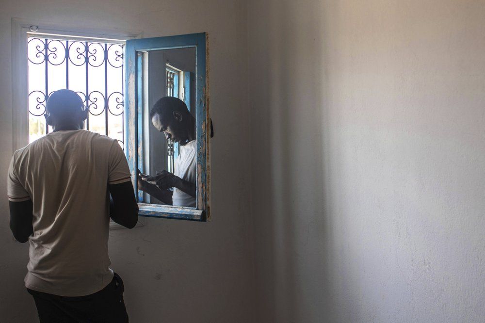 In this Monday, Sept. 23, 2019 photo, Abdullah, 25, a Sudanese migrant who tried crossing the Mediterranean from Libya, is reflected in a window as he uses his phone inside a Tunisian Red Crescent facility in Zarzis, southern Tunisia. The group of 47 in his first crossing from Tripoli over a year earlier had paid a uniformed Libyan and his cronies $127,000 in a mix of dollars, euros and Libyan dinars for the chance to leave their detention center and cross in two boats. They were intercepted in a coast guard boat by the same uniformed Libyan, shaken down for their cell phones and more money, and tossed back into detention. Image by Renata Brito/AP Photo. Libya, 2019.