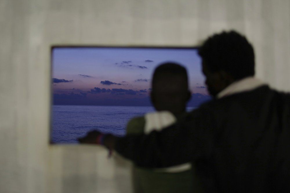 In this Sept. 13, 2019 photo, migrants rescued at sea look out the window aboard the Ocean Viking humanitarian ship as it sails in the Mediterranean Sea. The misery of migrants in Libya has spawned a thriving and highly lucrative business, in part funded by the EU and enabled by the United Nations, an Associated Press investigation has found. Image by Renata Brito/ AP Photo. Libya, 2019,