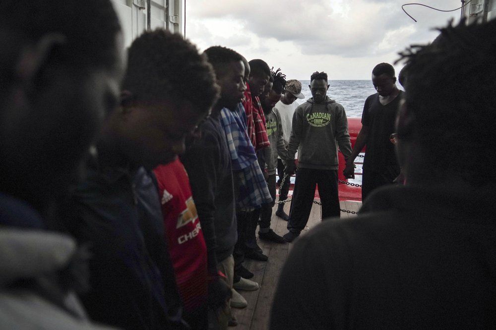 In this Sept. 21, 2019 photo, Eric Boakye, background center wearing a hoodie sweatshirt, and other rescued migrants pray for Europe to grant them a place of safety as they wait aboard the Ocean Viking humanitarian ship in the Mediterranean Sea. Boakye, a Ghanian, was locked in the al-Nasr Martyrs center in Libya twice, both times after he was intercepted at sea. The first time, his jailers simply took the money on him and set him free. He tried again to cross and was again picked up by the coast guard and returned to his jailers. Image by Renata Brito/AP Photo. Libya, 2019.
