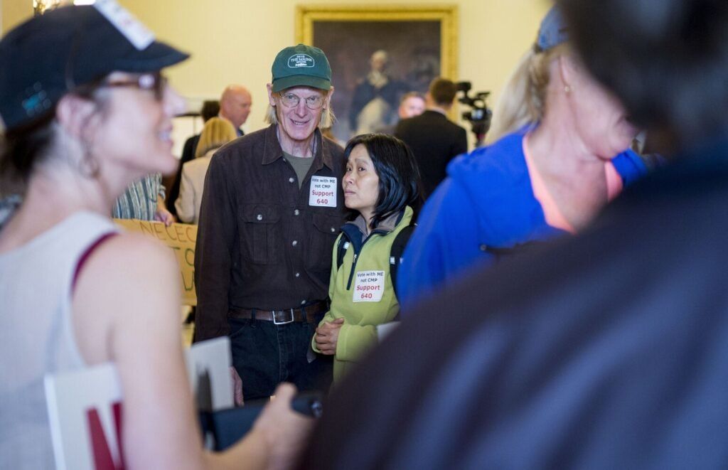 Duane Hanson and his partner Sally Kwan stand with others opposed to the New England Clean Energy Connect corridor project at the Maine State House in Augusta on June 4, 2019. Image by Michael G. Seamans. United States, 2019.