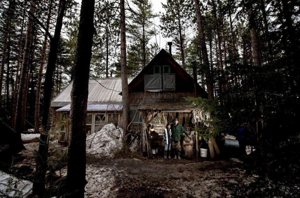 Sally Kwan and Duane Hanson pose for a portrait at their summer kitchen outside their cabin on Whipple Pond on April 23, 2019. The couple who power their cabin from a wood stove and two solar panels worry they'll be able to see the 100-foot tall transmission line above the white pine trees that surround their property. Image by Michael G. Seamans. United States, 2019.
