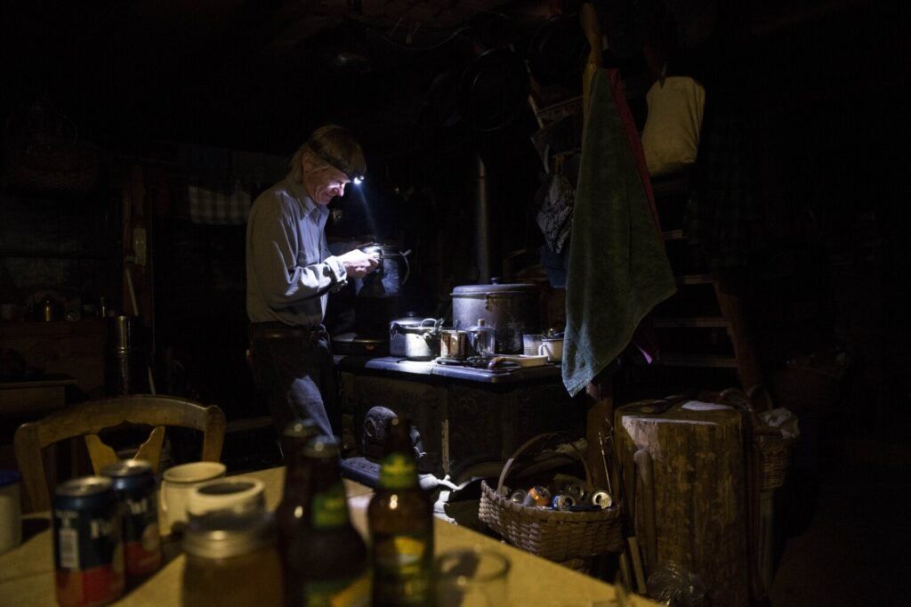 T5 R7, MAINE - MAY 26, 2019 Duane Hanson prepares dinner over his wood-fired stove at his homestead in the Unorganized Territories in the north woods of Maine near T5 R7 on May 26, 2019. Hanson has two solar panels and one light. Cooking over the stove by headlamp is not uncommon. Image by Michael G. Seamans. United States, 2019.