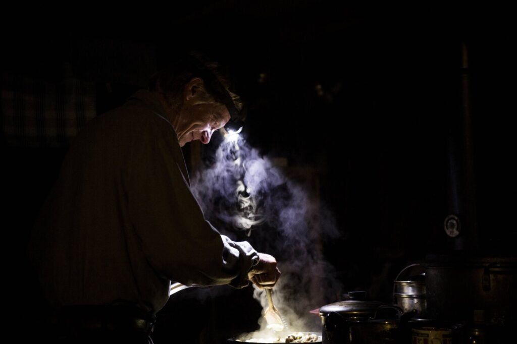 Duane Hanson prepares dinner over his wood-fired stove at his homestead in the Unorganized Territories in the north woods of Maine near T5 R7 on May 26, 2019. Hanson has two solar panels and one light. Cooking over the stove by headlamp is not uncommon. Image by Michael G. Seamans. United States, 2019.