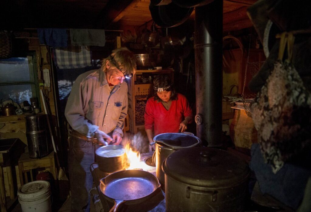 Duane Hanson and Sally Kwan prepare dinner at their home on Whipple Pond in T5 R7 in the Unorganized Territories of Maine on May 26, 2019. Using an antique wood stove, they cook dinner and heat the cabin. Image by Michael G. Seamans. United States, 2019.