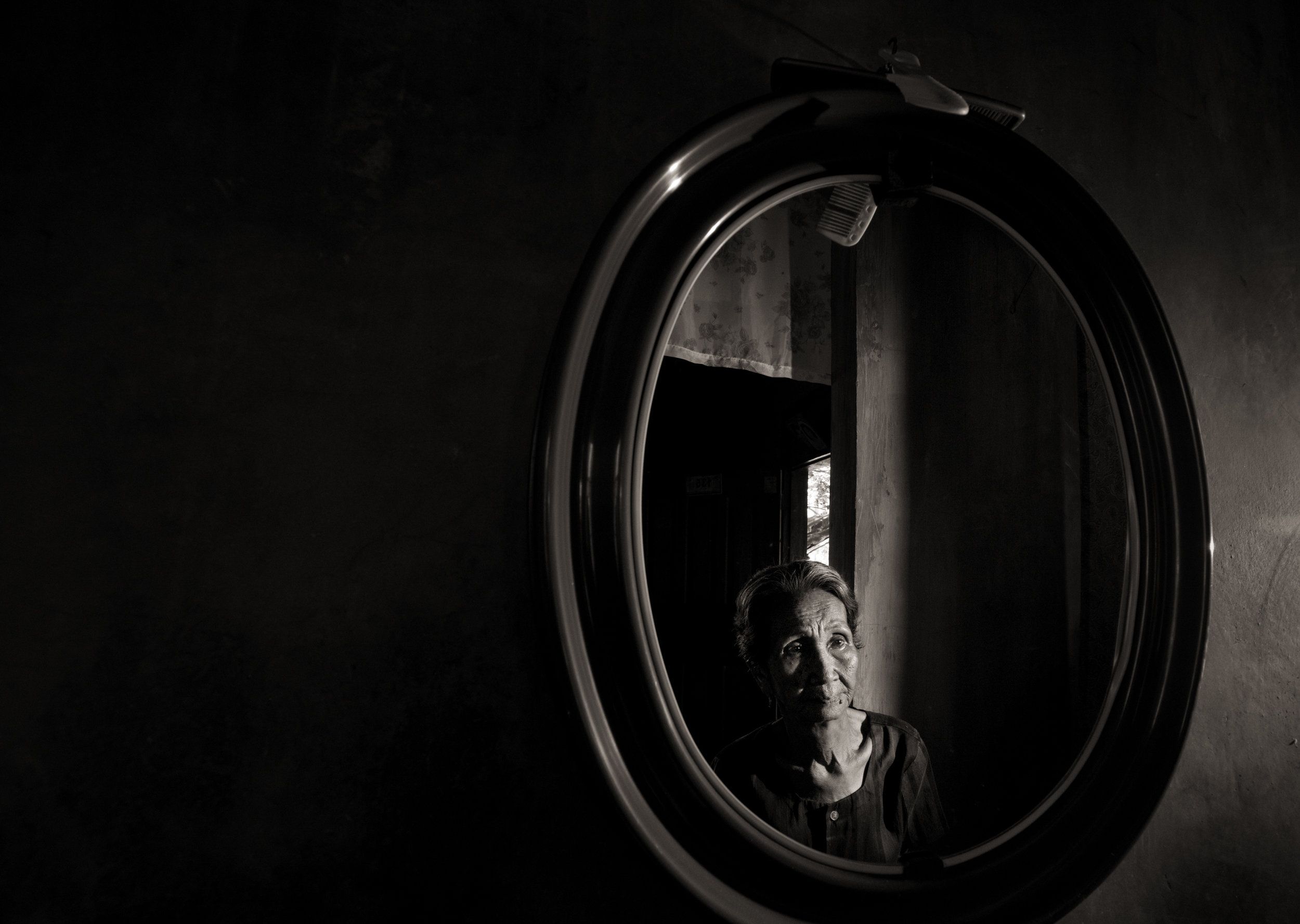Emilia dela Cruz Mangilit is reflected in a mirror in her home in the village of Mapaniqui. Mangilit was only 15 years old when her village was shelled, then attacked by the Japanese during World War II. Image by Cheryl Diaz Meyer. Philippines, 2019.