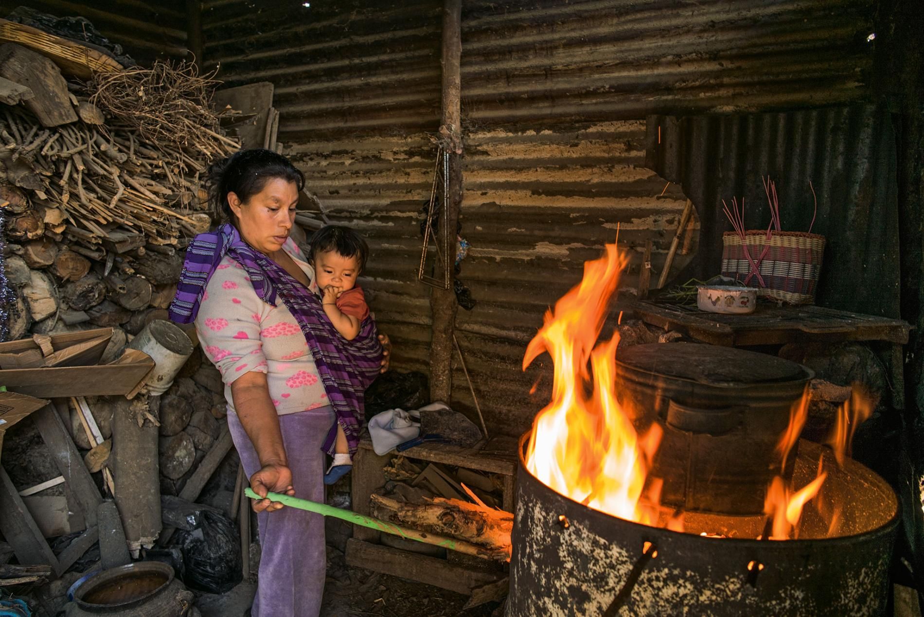 Near Antigua Guatemala, eight-month-old Pablito keeps an eye on breakfast as his mother, Angélica Epatal Garcia, tends to the makeshift barrel stove. She and her daughters walk 45 minutes each way to collect the wood for three daily fires. Image by Lynn Johnson. Guatemala, 2017.