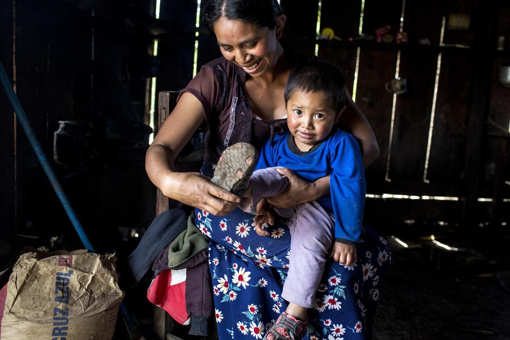 Catarina Domingo, dressing her son, lives in a house that is one of the few remaining in Yalambojoch that is made of wood and has a tin roof and a dirt floor. Image by Simone Dalmasso. Guatemala, 2019. 

