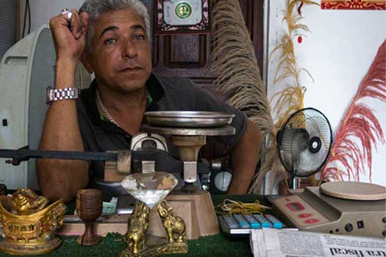 A Tumeremo gold pawn shop owner sits behind his desk while enjoying the luxury of a cooling desk fan. Image by Bram Ebus. Venezuela, 2017.
