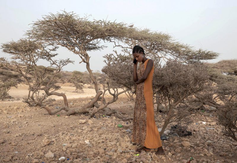 Hades, 16, an Ethiopian Tigray migrant, makes a phone call to her mother in Ethiopia as she takes shelter under trees at the last stop of her journey before leaving by boat to Yemen in the evening, in Obock, Djibouti, July 15, 2019. Hades did not know that there is a civil war in Yemen. She was told by her guide that women are safe in homes of their employers in Saudi Arabia and that she would not face deportation. Image by AP Photo / Nariman El-Mofty. Djibouti, 2019.