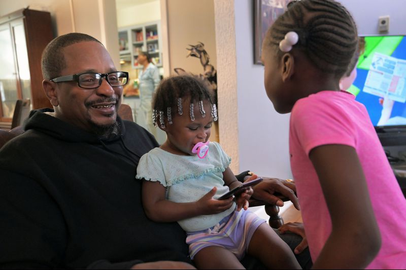 Cecil Burton rocks his granddaughters Chasity Shufford, 1, and Katelyn Ellis, 6, during a family gathering while their mother, Keisha, cooks fried chicken in the kitchen. Burton, 54, said child support once garnished so much of his paycheck he didn't have enough to live on. His coworkers took up a collection for him. Image by Karl Merton Ferron / Baltimore Sun. United States, 2019. 
