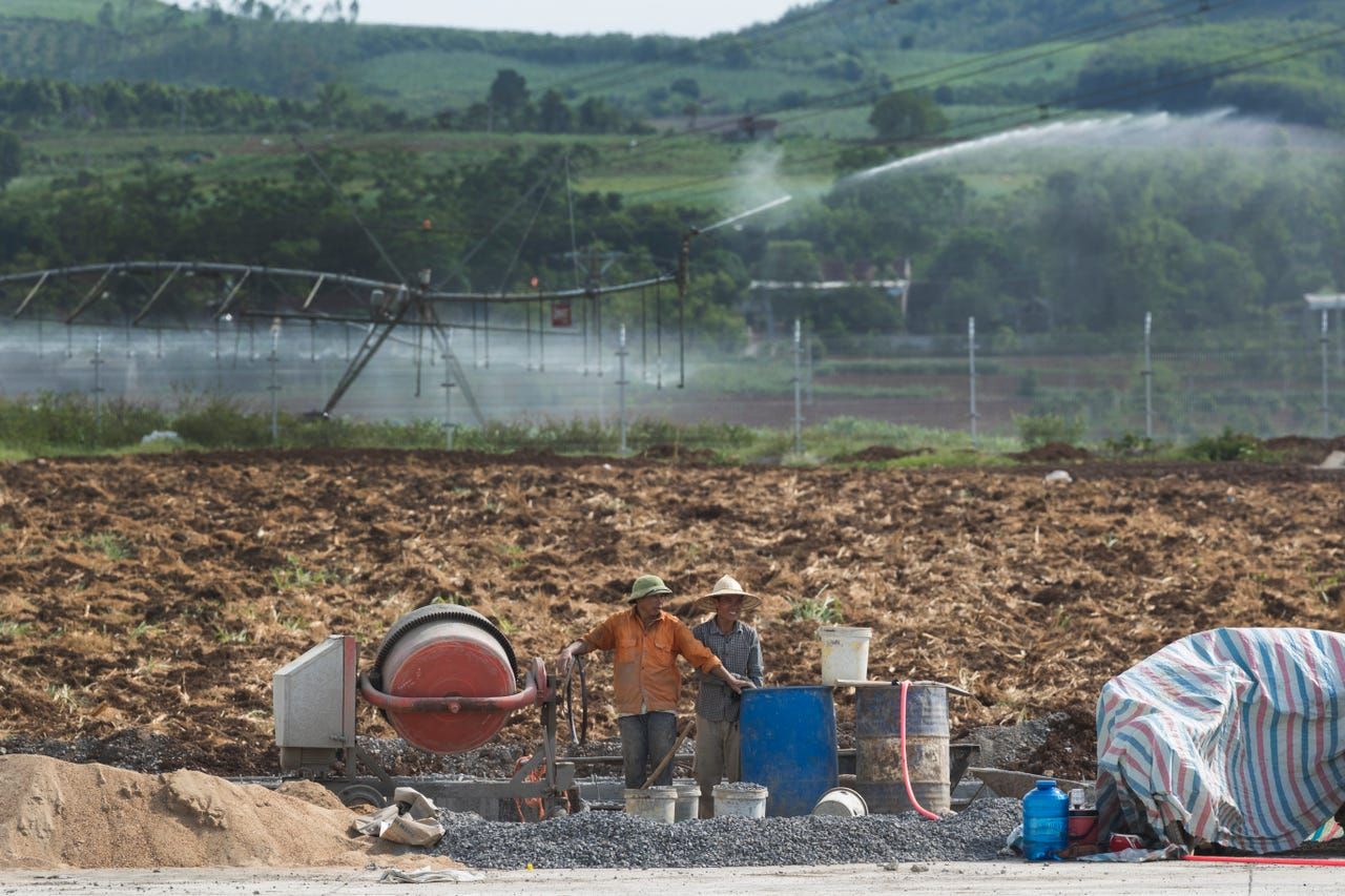 An irrigation system waters crops while construction workers build a parking lot at the feed warehouse at TH Milk's operations in Nghia Son, Vietnam. Without irrigation, a field can produce one harvest of corn annually. With irrigation, corn can be harvested three times per year. Image by Mark Hoffman. Vietnam, 2019. 
