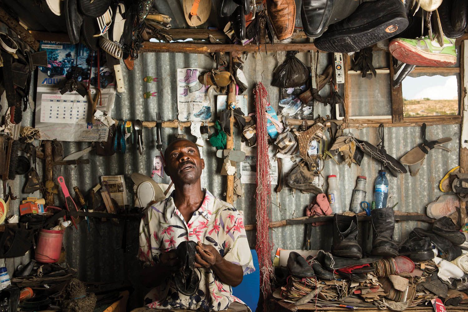 Eddy Bien Aime fixes a pair of sandals at his business in the neighborhood known as Canaan 2. Most of his business involves selling footwear he has bought secondhandand refashioned. He moved to Canaan just days after the earthquake and claimed a large parcel of land, some of which he has since given away. Image by Allison Shelley. Haiti, 2017.
