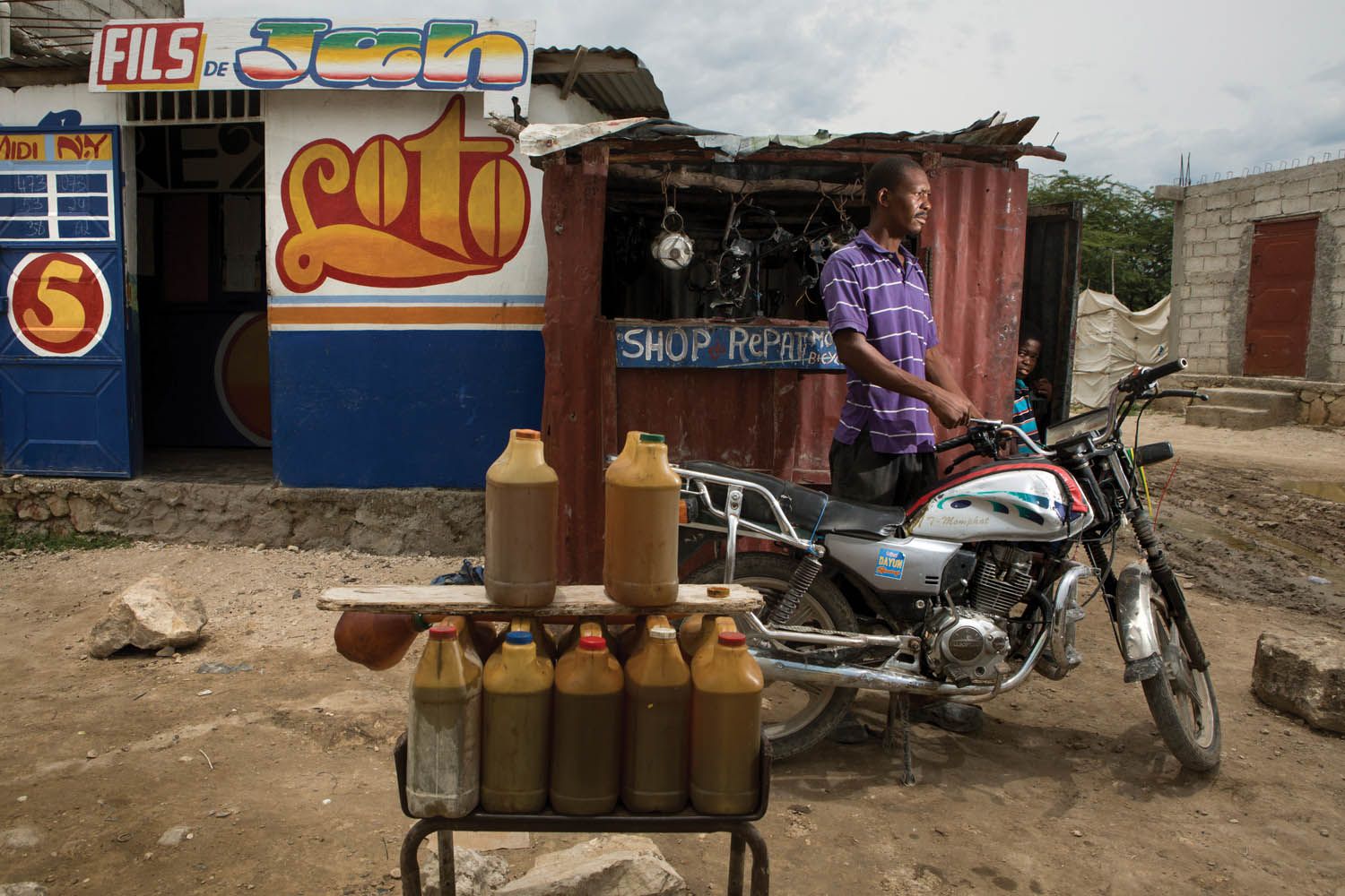Jacob Viknel at his motorcycle-repair shop in the Jerusalem section of Canaan. When customers pull up, Viknel fills their tanks with gasoline from the jugs on display. Image by Allison Shelley. Haiti, 2017.