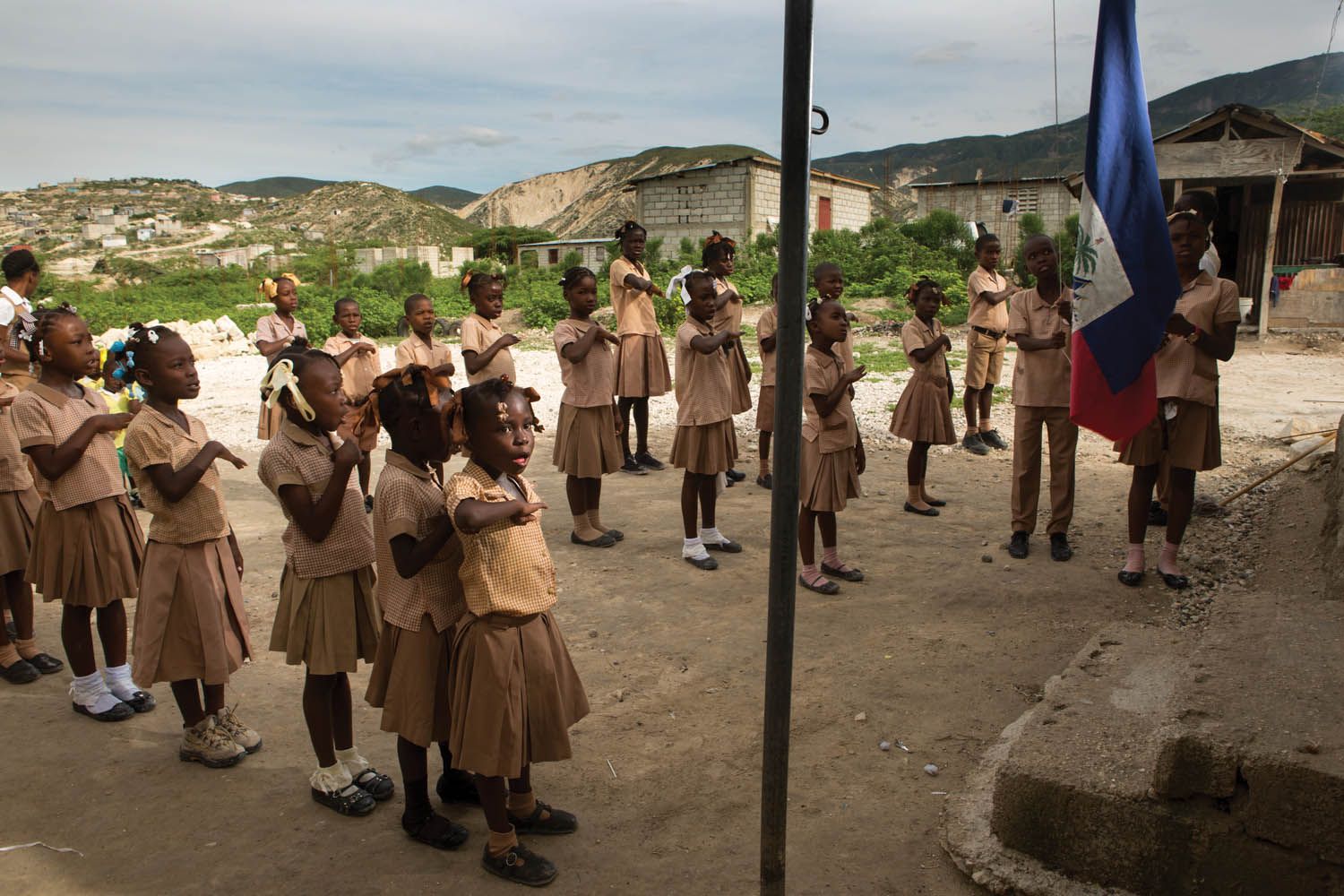Children sing the national anthem as the Haitian flag is raised at their elementary school, which doubles as the Church of the Nazarene, in the Onaville neighborhood. Pastor Marc Loumette founded the church and school in 2010, after his calling to minster to those affected by the earthquake. Image by Allison Shelley. Haiti, 2017.