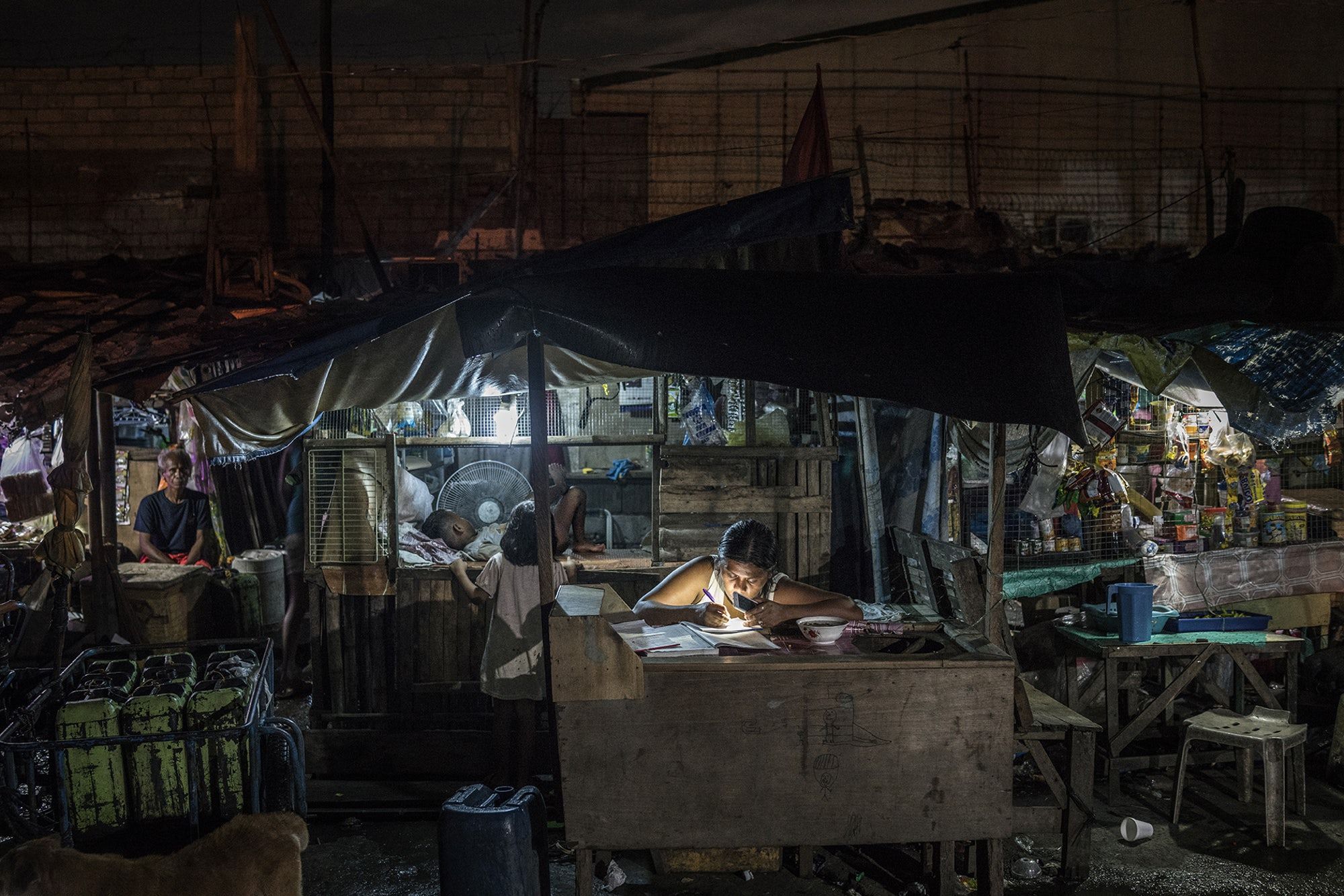 A woman works on bookkeeping using the light of her phone in the Market 3 slum in the Navotas Fish Port Complex. Market 3 has electricity but no plumbing. Residents often turn to drugs to alleviate hunger and stay awake for the long hours they work at low pay. Image by James Whitlow Delano. Philippines, 2017.