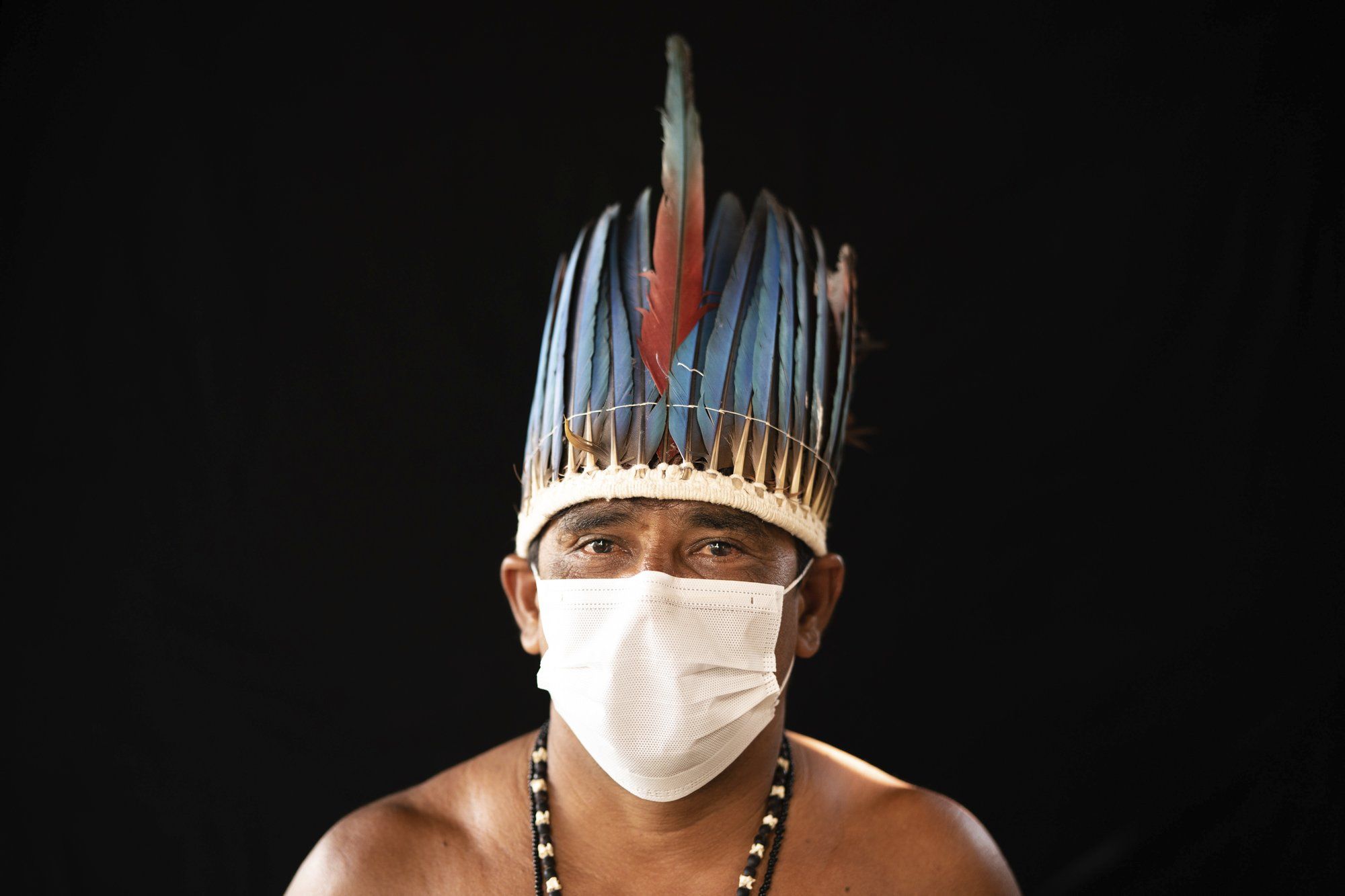Eleomar da Silva, 44, of the Sateré Mawé indigenous ethnic group, poses for a portrait wearing the traditional dress of his tribe and a face mask amid the spread of the new coronavirus in the Gaviao community near Manaus, Brazil, Friday, May 29, 2020. Image by Felipe Dana / AP Photo. Brazil, 2020.