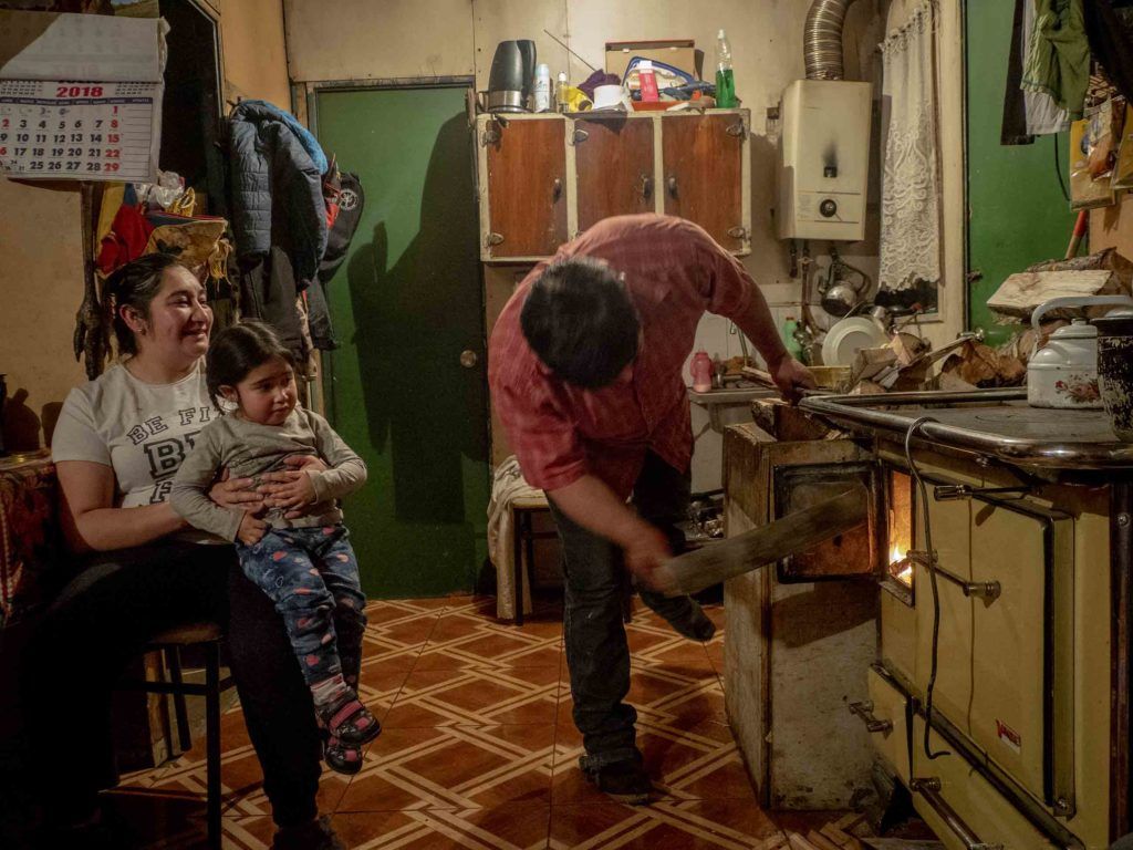 At home, Marcelo fires up his own stove to heat his tiny home, while his wife Jessica Vera Vidal and their daughter, Jazmine Aborto Vera, 3, watch. Image by Larry C. Price. Chile, 2018.