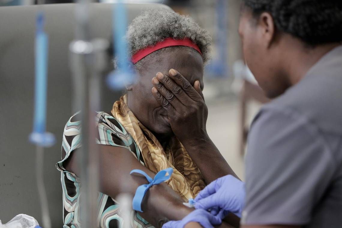 A cancer patient at the University Hospital of Mirebalais covers her face as a nurse prepares her to receive chemotherapy. Image by José A. Iglesias. Haiti, 2018.