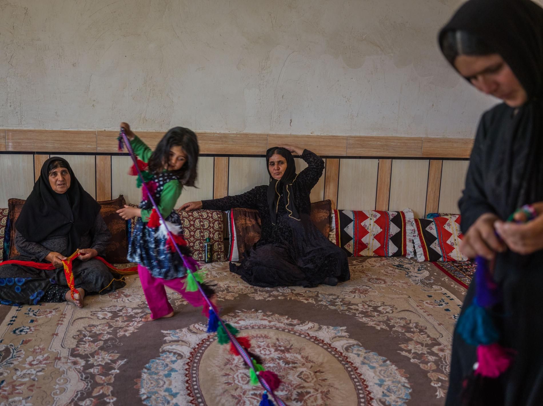 Bibi Jan Mokhtari (center), 57, and Mahpasand Mokhtari (left), 62, are living in a house for the first time. Elham Mokhtari, six, plays with a handicraft made by her mother, Mahnaz Mokhtari (far right), 39. The family makes and sells handicrafts as a main source of income. Image by Newsha Tavakolian. Iran, 2018.