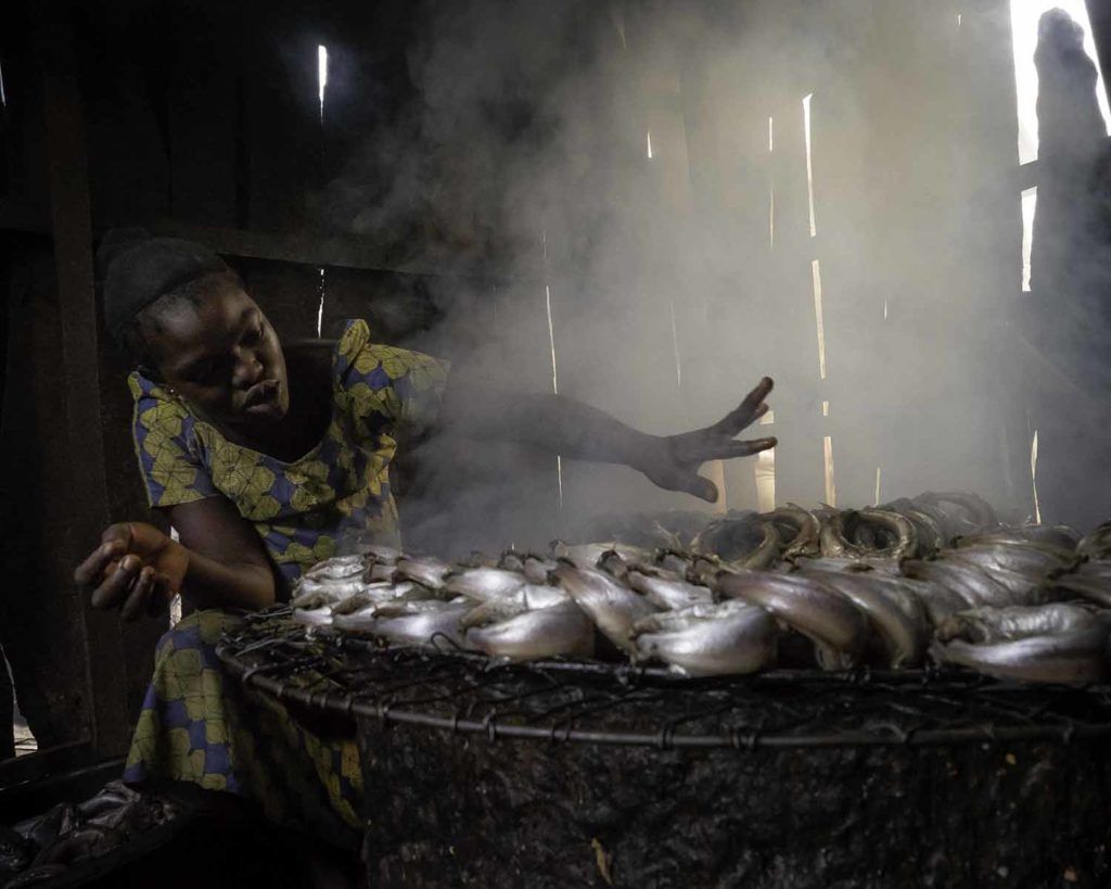 The air grows thick with early-morning cooking fires. Here in the impoverished Makoko neighborhood of Lagos, a woman is smoking fish — a ritual repeated in thousands of other homes. Image by Larry C. Price. Nigeria, 2018.