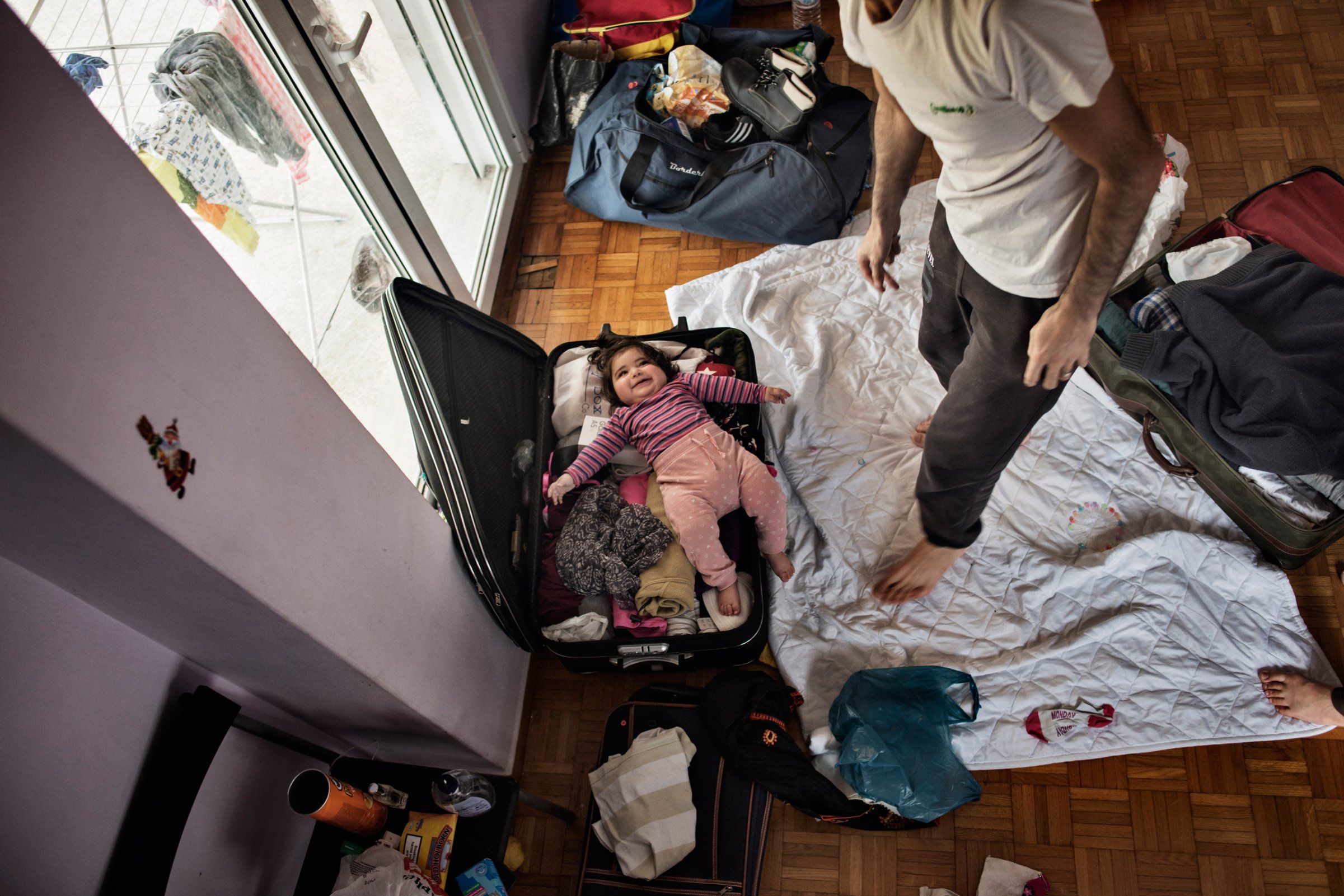 Syrian refugee Muhanned Abzali, jokes with his 6 month old daughter, Heln, by packing her in a suitcase, the night before leaving for relocation in Estonia. Image by Lynsey Addario. Greece, 2017. 